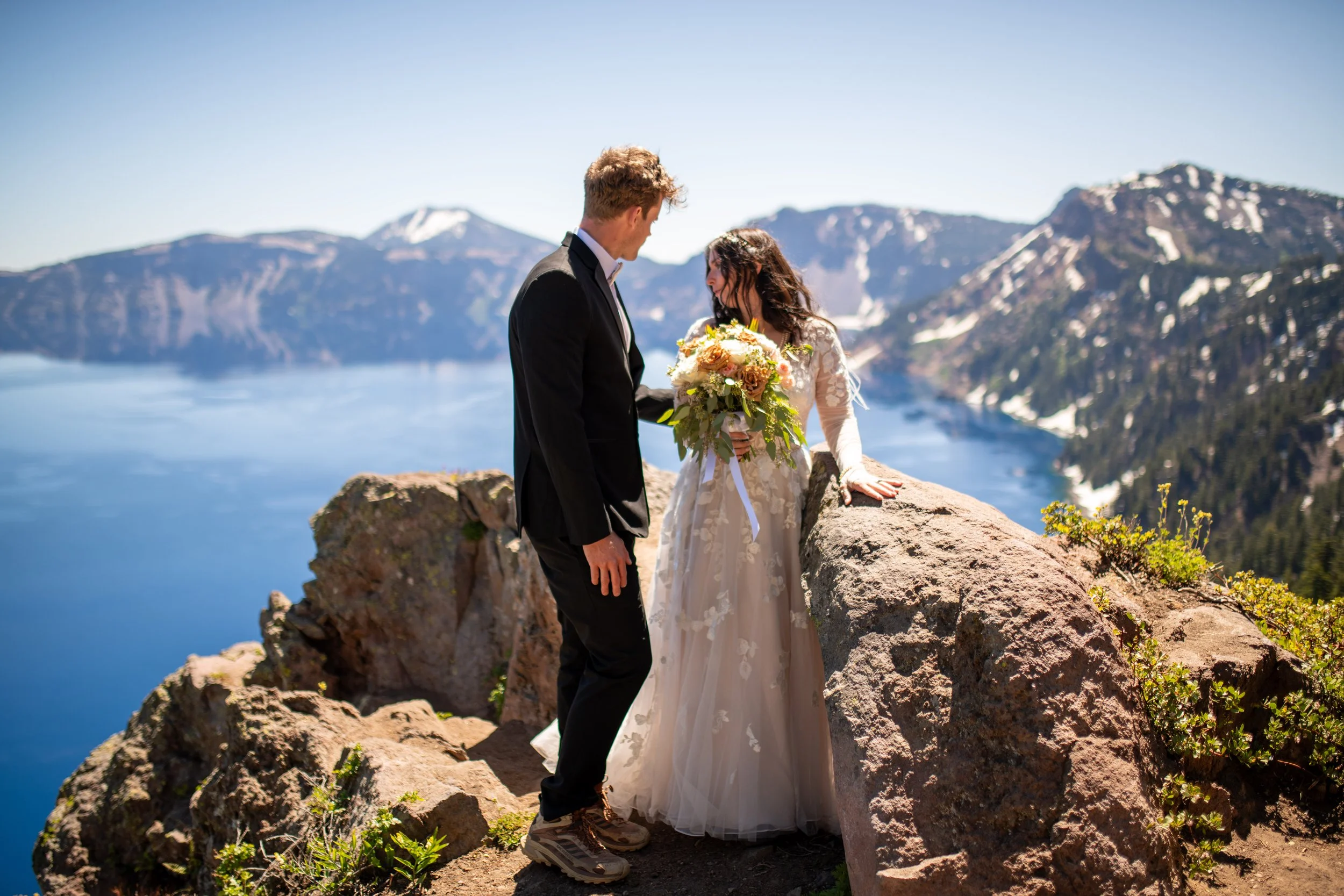 A couple in wedding attire stands on rocks overlooking a lake and mountain range under clear blue skies.