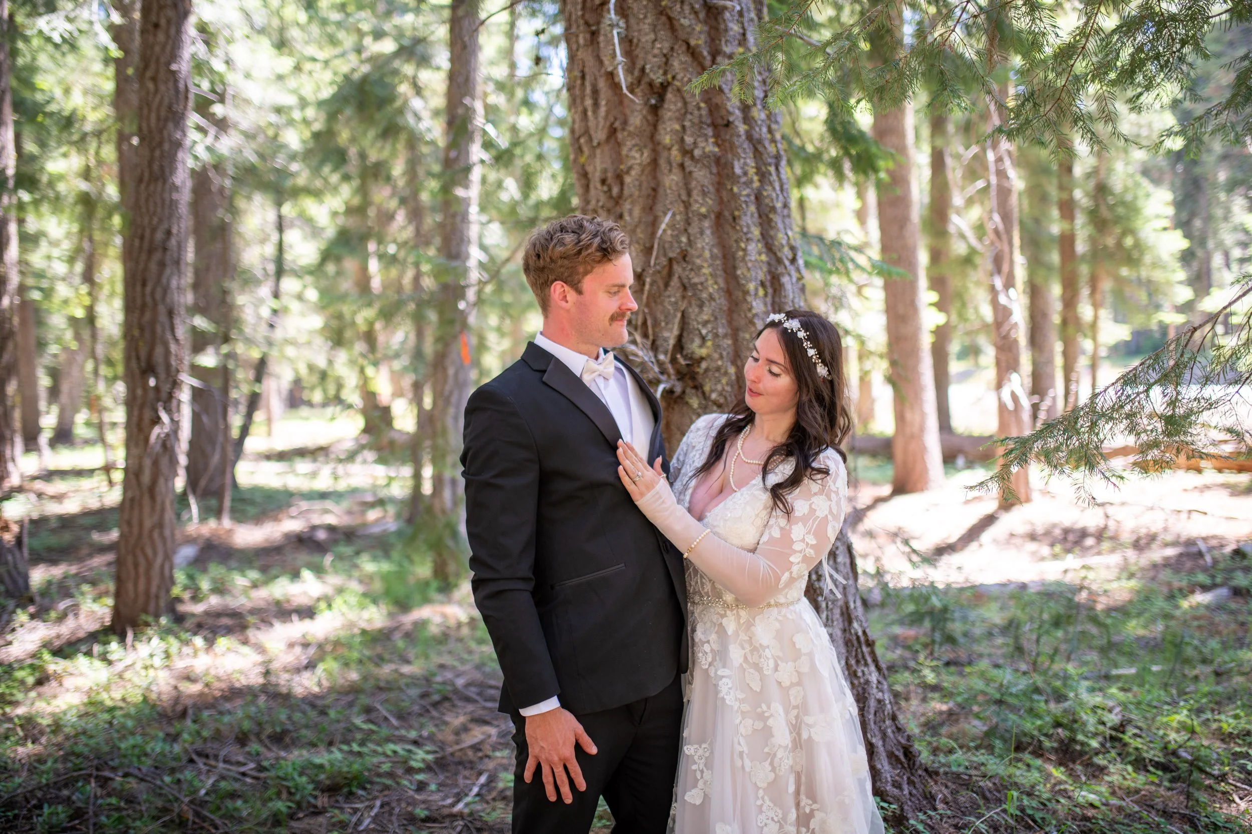 A bride and groom standing close together outdoors in a forest, with the bride touching the groom's chest. The groom is wearing a black tuxedo with a white shirt and bow tie. The bride is wearing a white lace wedding dress with gloves and a floral he