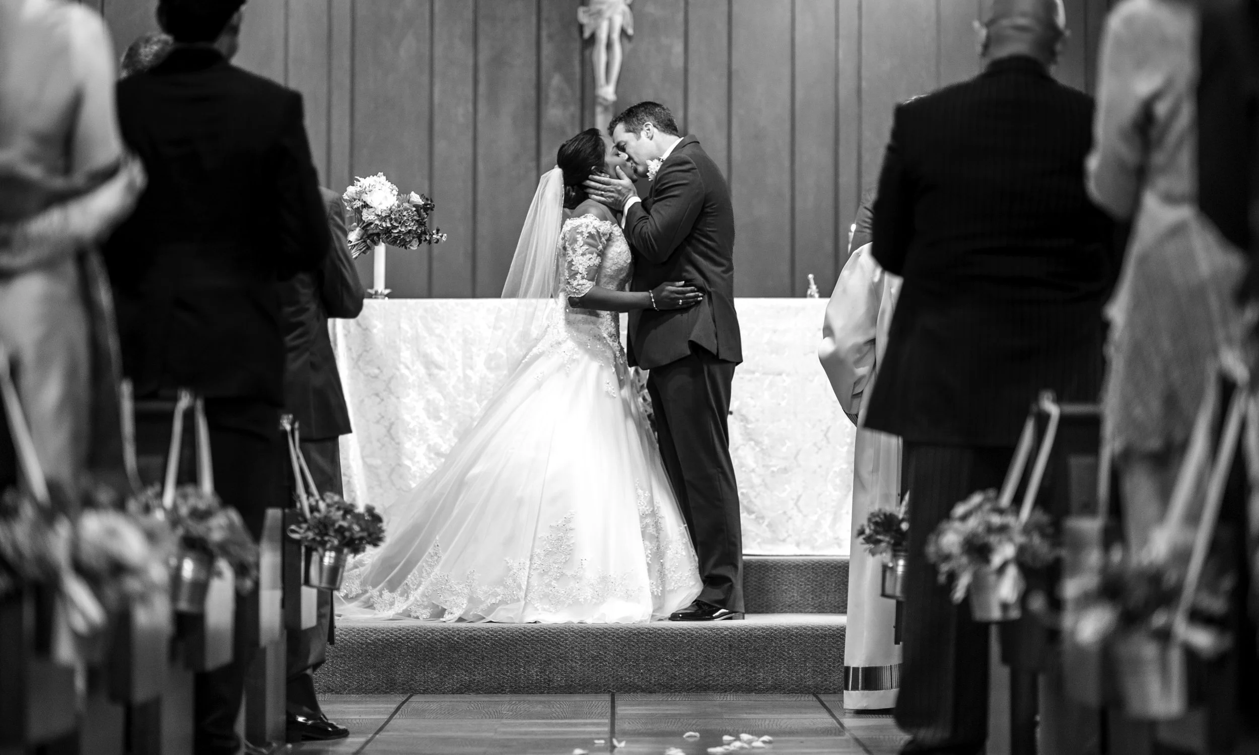 A black and white photo of a wedding ceremony where a bride and groom are kissing at the altar. The bride is in a wedding dress and veil, and the groom is in a suit. Guests are standing on either side, watching the couple.