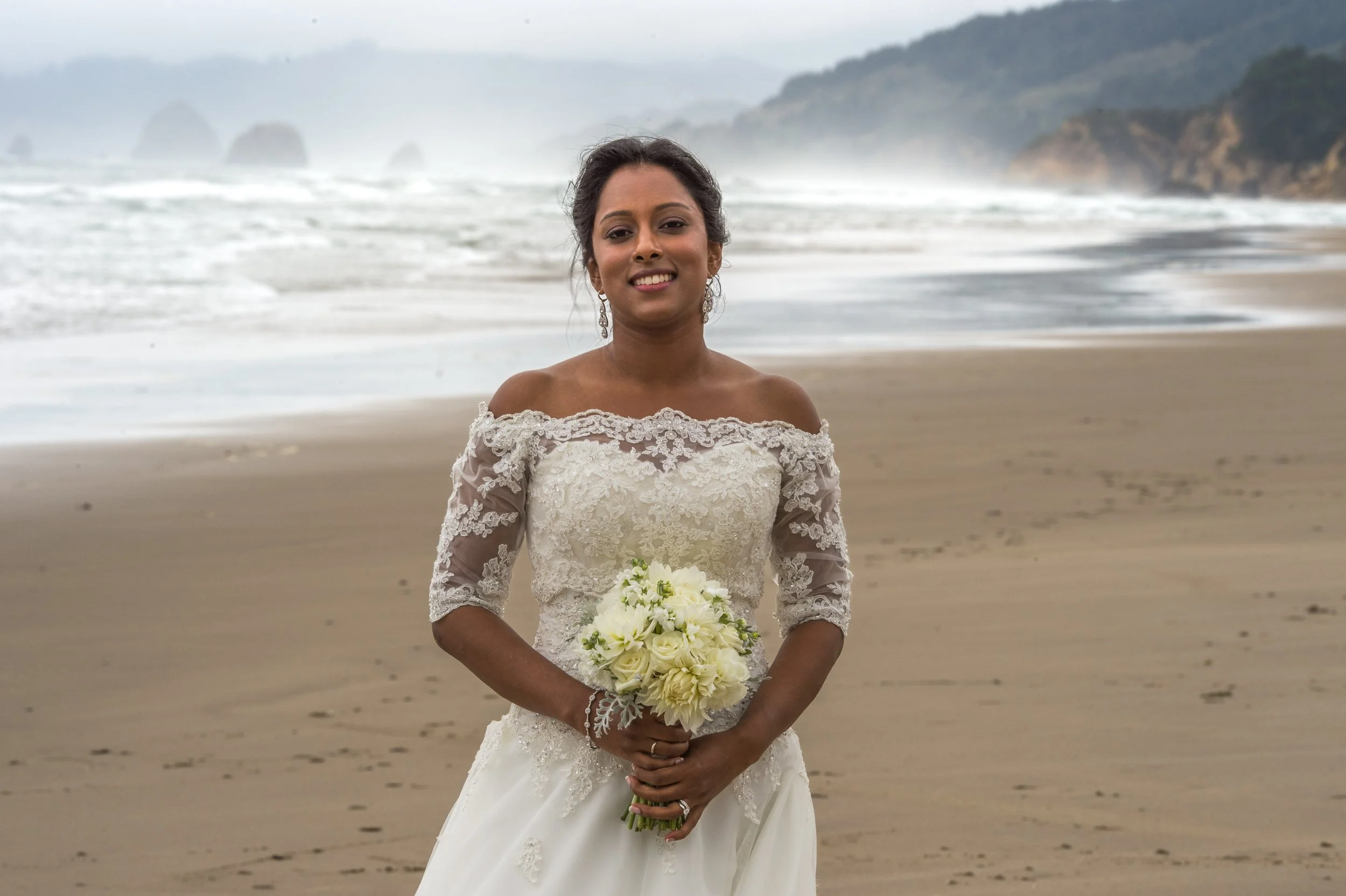 A woman in a wedding dress holding a bouquet of white flowers, standing on a sandy beach with ocean waves and rocky cliffs in the background.