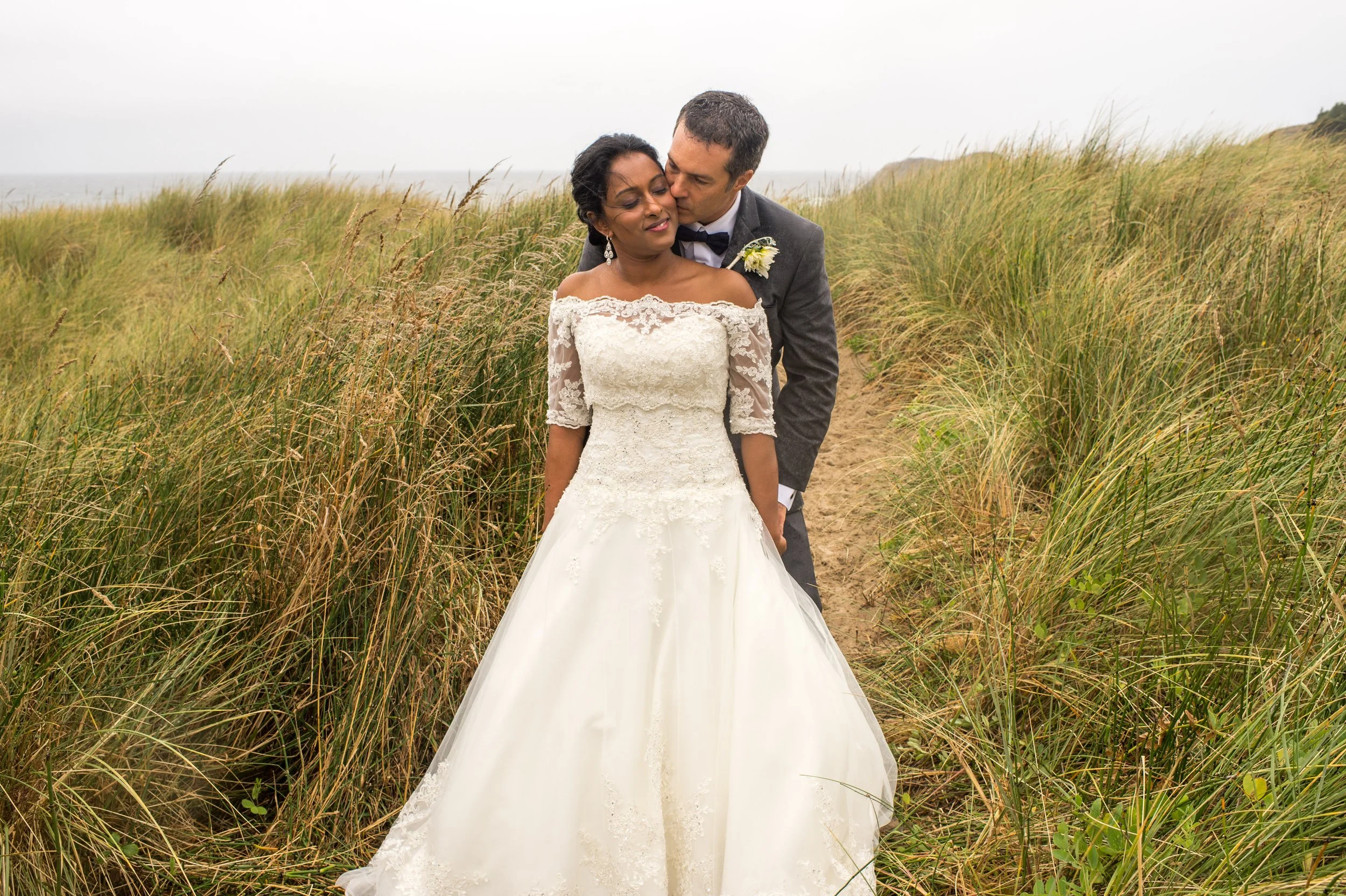A bride and groom stand together on a sandy path surrounded by tall grass in an outdoor setting, with the groom kissing the bride on the side of her head.
