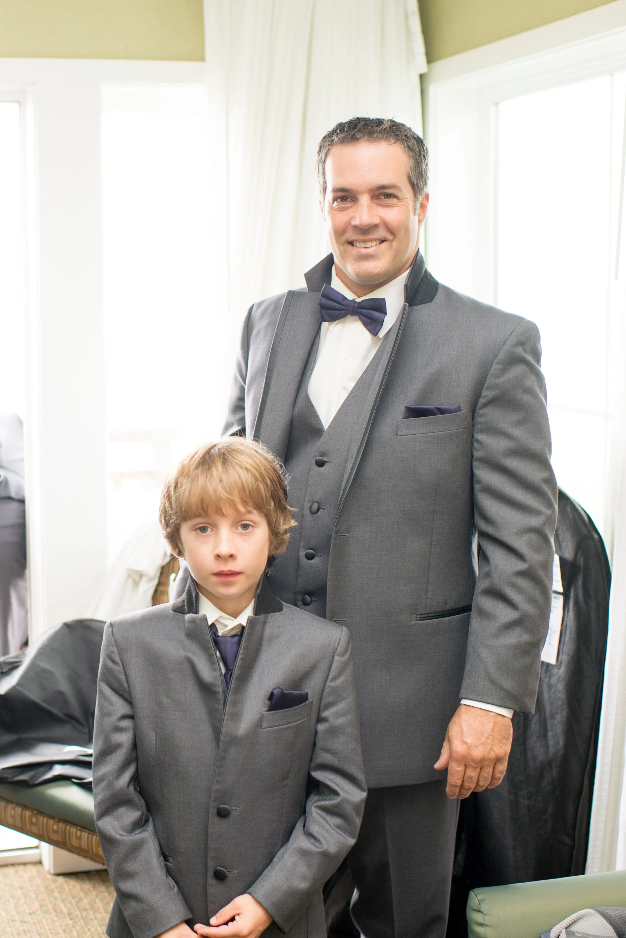A man and a boy dressed in matching grey tuxedos and bow ties, standing in a room with large windows and white curtains, possibly preparing for a formal event.