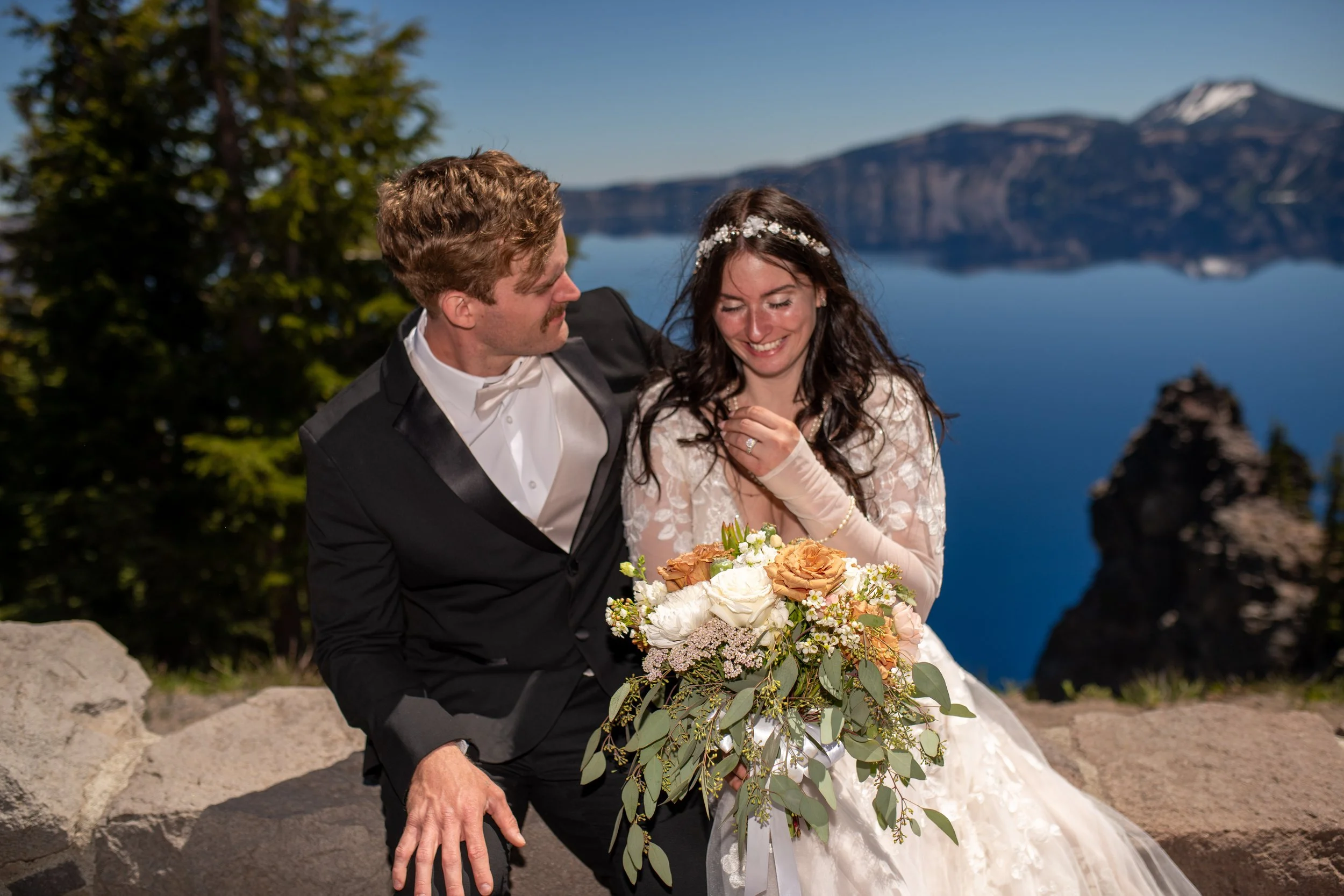 A smiling bride holding a bouquet of white and peach roses and greenery, standing next to a groom in a tuxedo with a mountain and lake in the background.