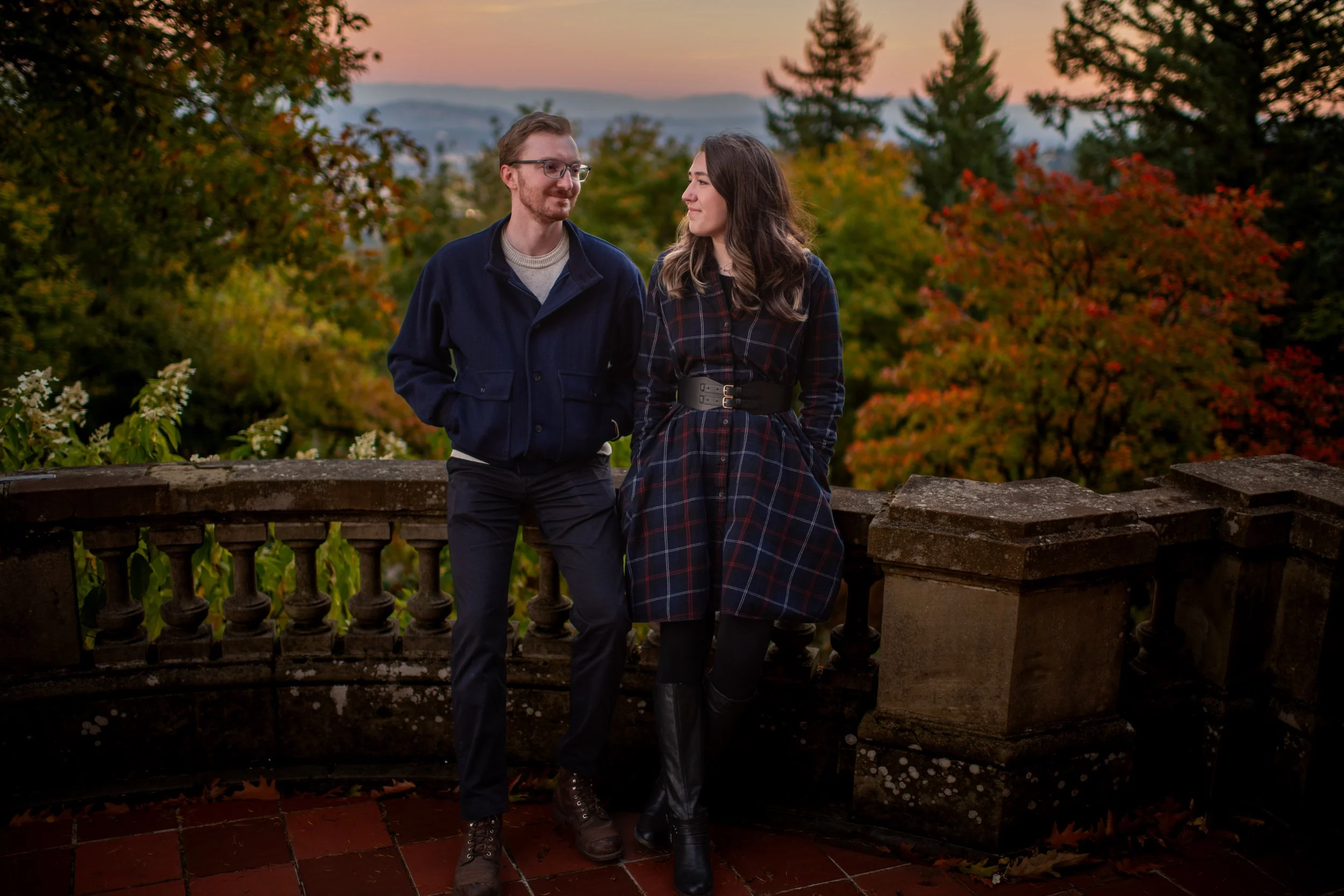 A young man and woman standing outdoors on a stone balcony during sunset, surrounded by autumn-colored trees.