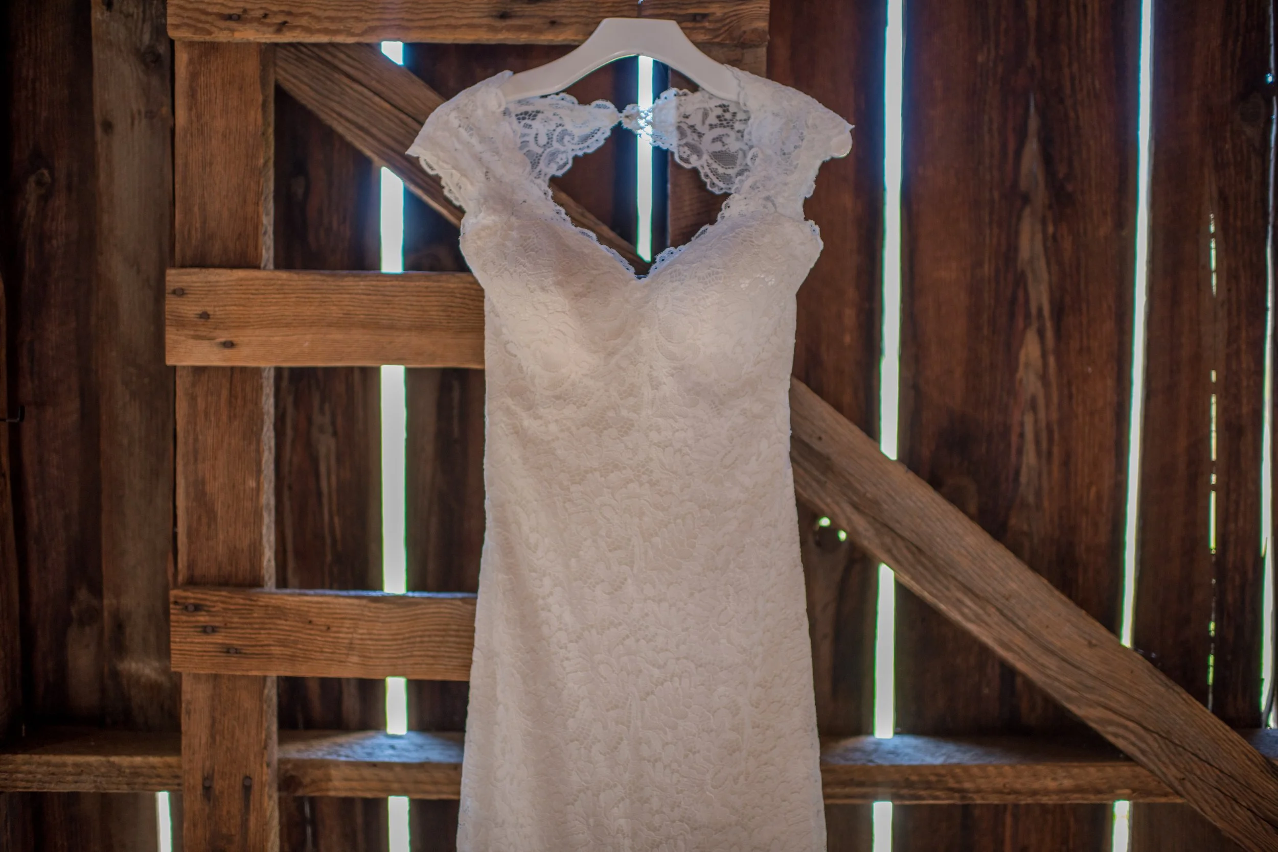 A white lace wedding dress hanging on a white hanger against a wooden barn wall with gaps allowing light to shine through.