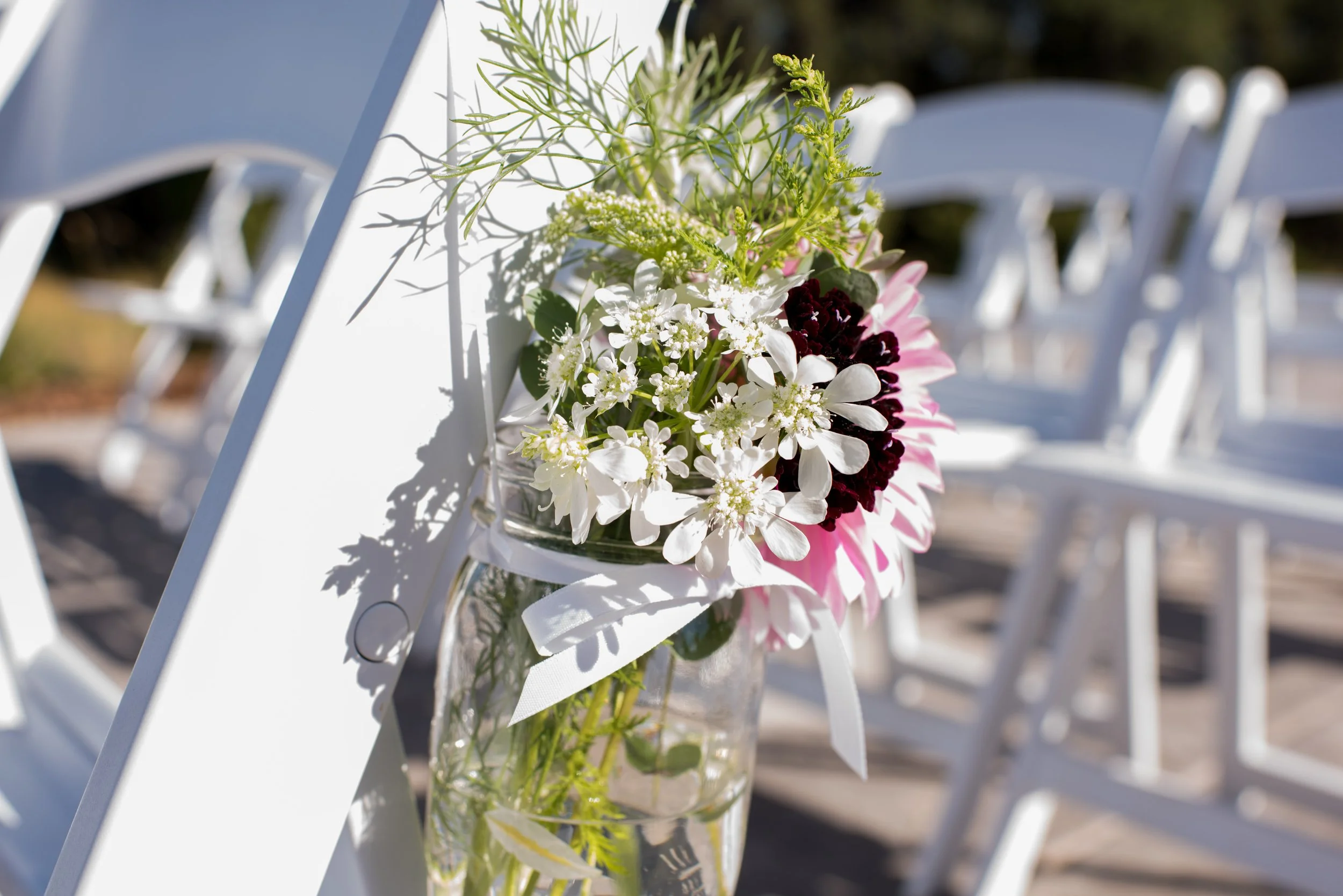 A glass jar filled with various flowers including white, pink, and dark red, attached to a white chair at an outdoor event.
