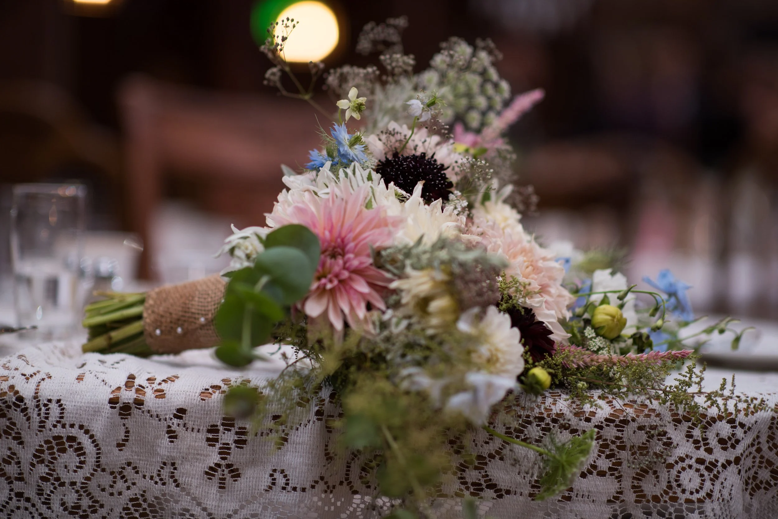 Close-up of a floral arrangement on a lace tablecloth, featuring pink, white, and blue flowers with green leaves, and wrapped in burlap, with a blurred dark background and soft lighting.