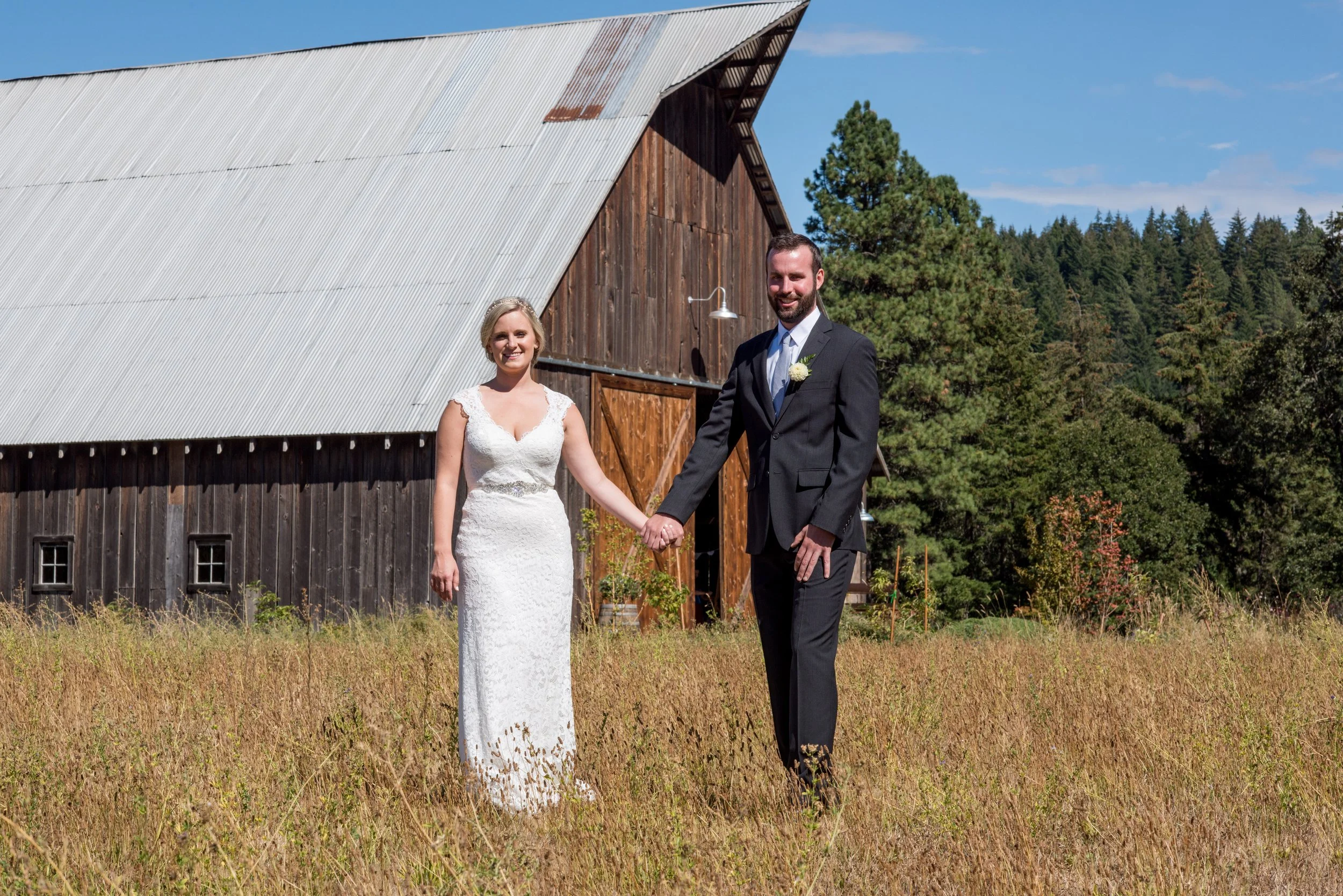 A bride in a white wedding dress and a groom in a black suit holding hands outdoors in front of a rustic barn and green trees on a sunny day.