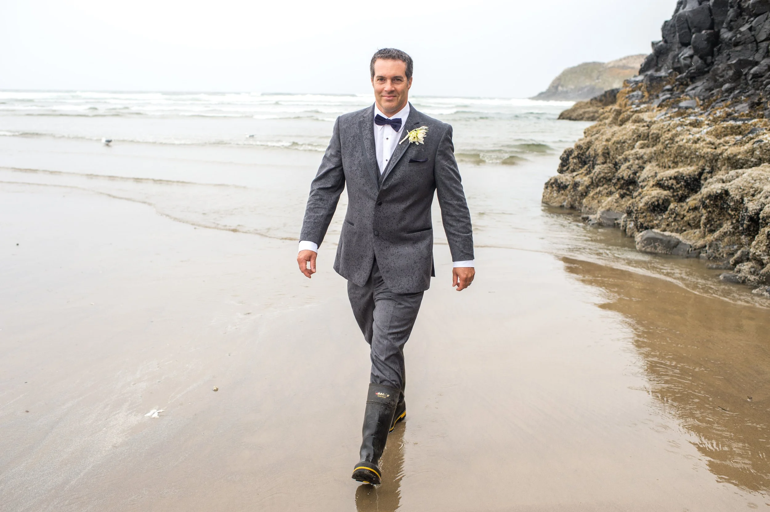 A man dressed in a tuxedo with a bow tie and boutonnière walking along a beach with rocky cliffs in the background, while wearing rain boots.