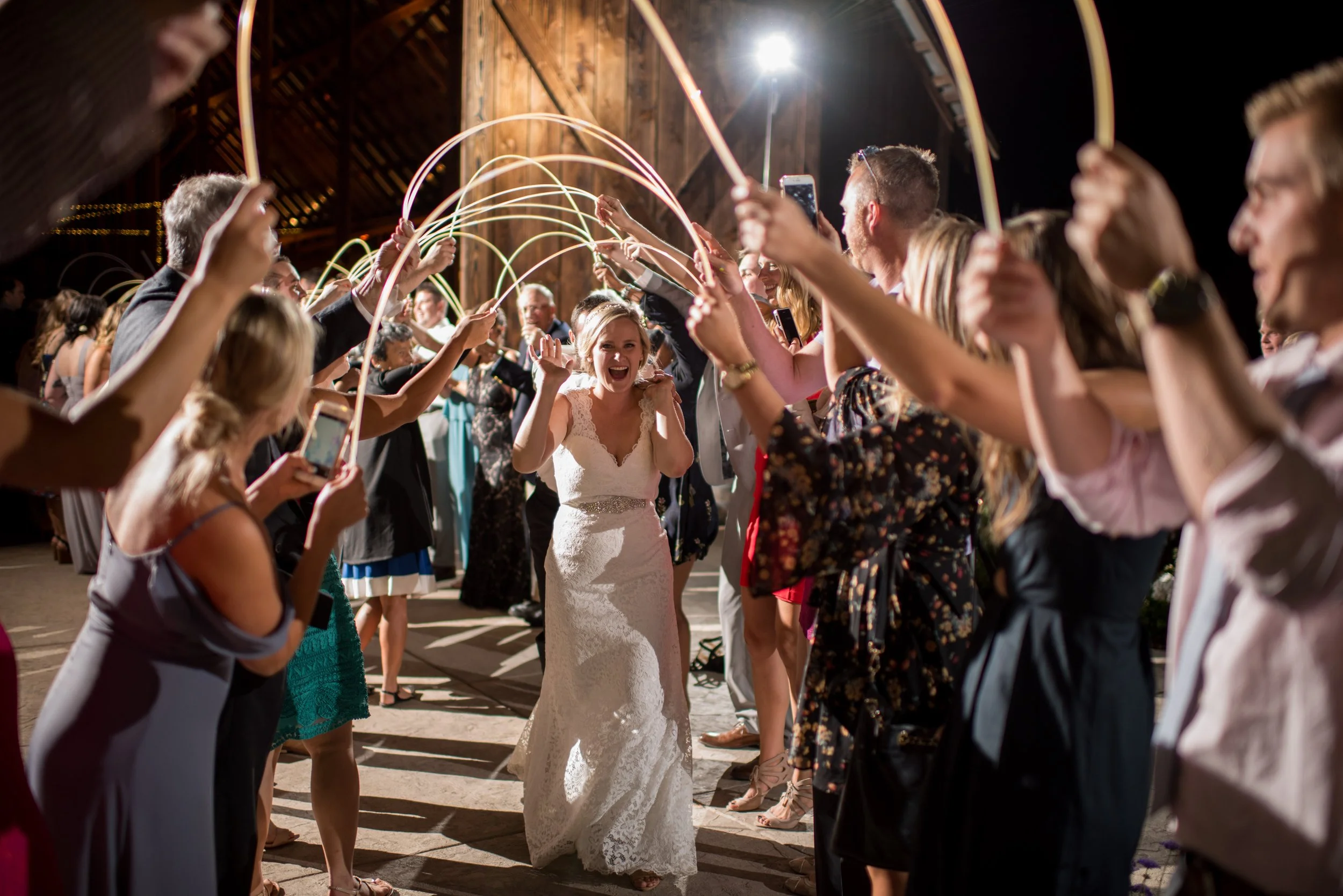 Group of people surrounding a bride at a wedding reception, holding glow sticks and taking photos inside a rustic barn.