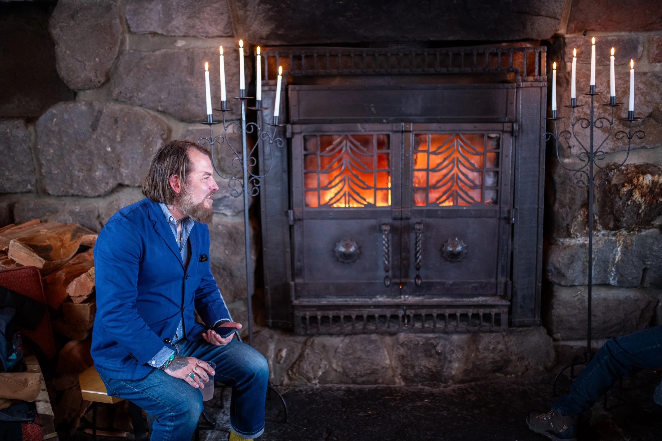 A man with a beard and long hair, wearing a blue jacket, sitting on a wooden bench. He is holding a cup and looking to the left. Behind him is a stone wall and a large metal fireplace with glowing fire inside. There are two black metal candelabras wi