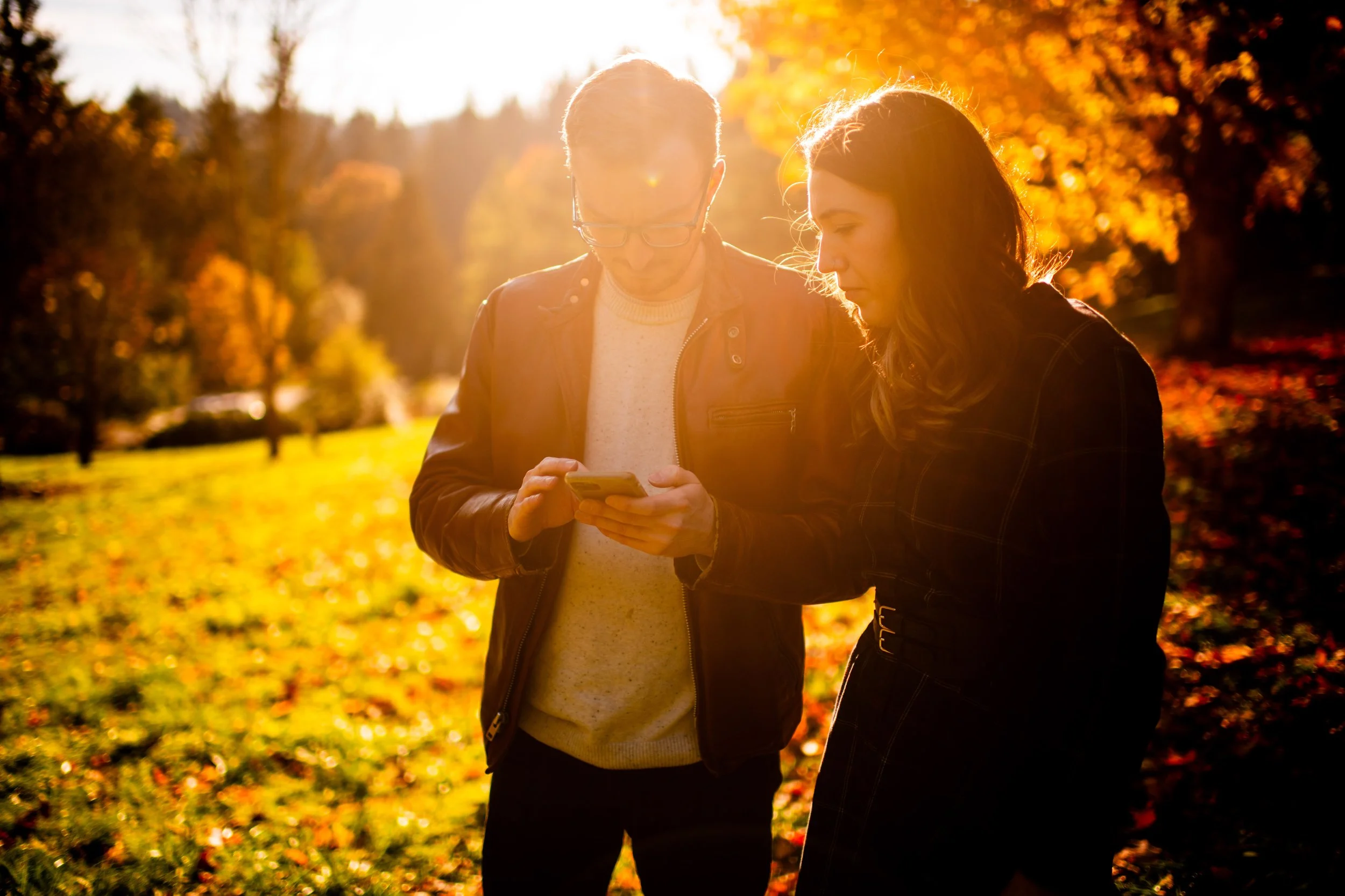 A man and woman standing outdoors in autumn, looking at a phone, with sunlight behind them and colorful fall trees in the background.