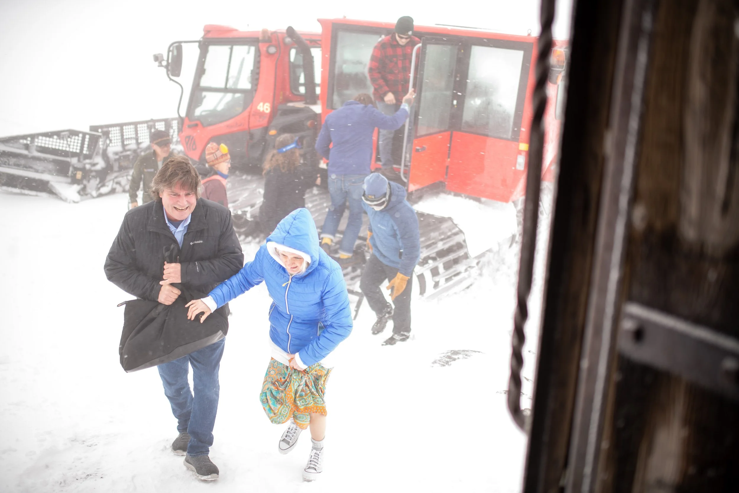 People exiting a red snowcat vehicle in snowy weather, some dressed in winter clothing, as seen through a window with a blurred wooden frame.