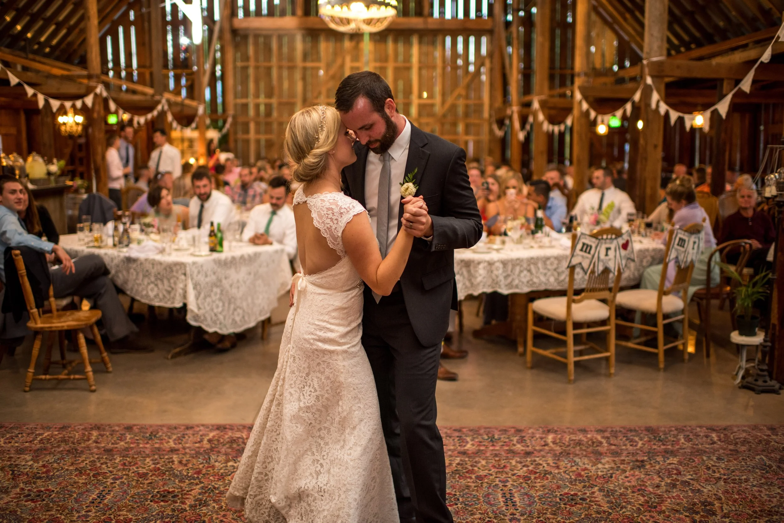 A bride and groom sharing their first dance at a wedding reception in a rustic barn decorated with string lights and bunting.
