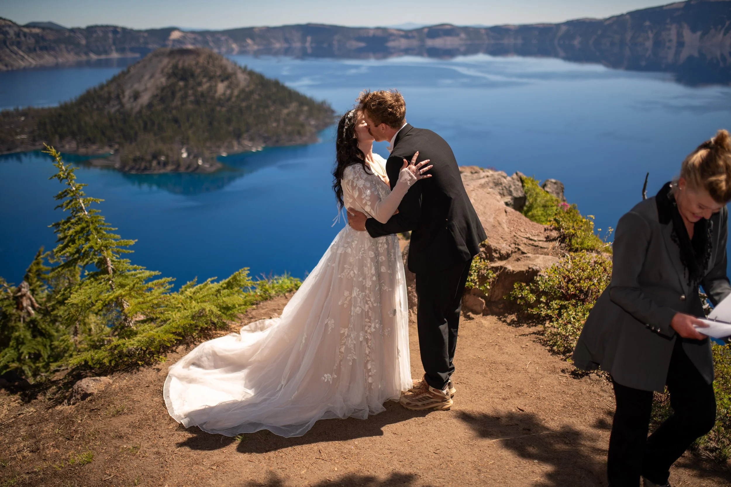 A bride and groom sharing a kiss at an outdoor wedding ceremony near a large blue lake surrounded by mountains. The bride is wearing a white lace wedding dress and the groom a black tuxedo. A woman with glasses is seen in the foreground, smiling and 