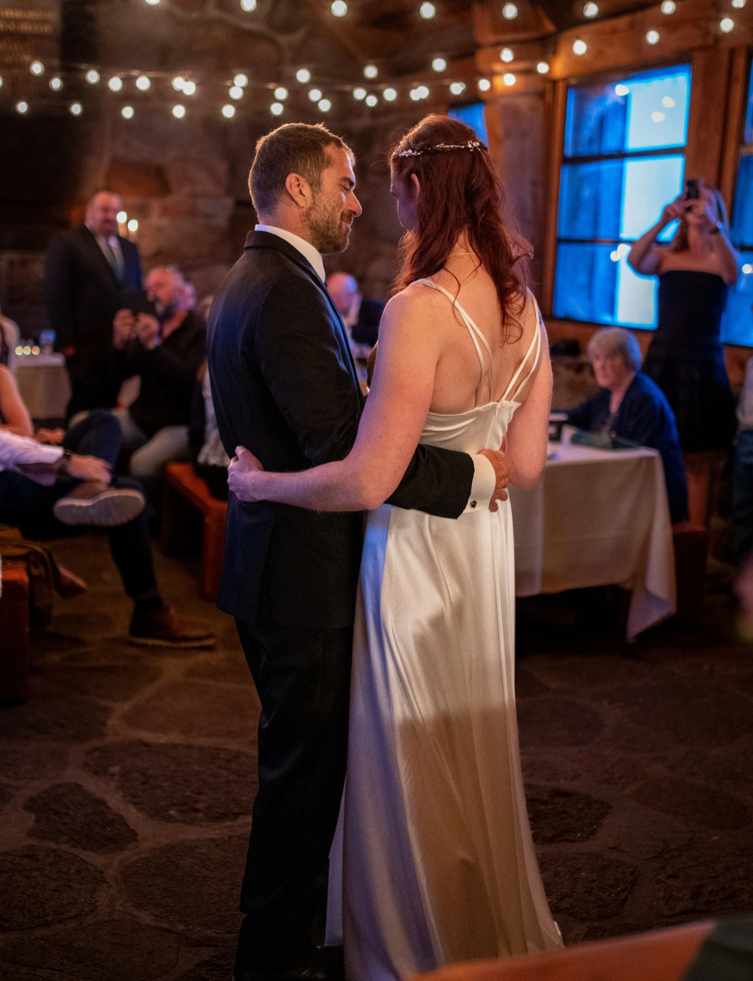 A couple sharing a dance at a wedding reception in a rustic barn with string lights and blue-lit windows.