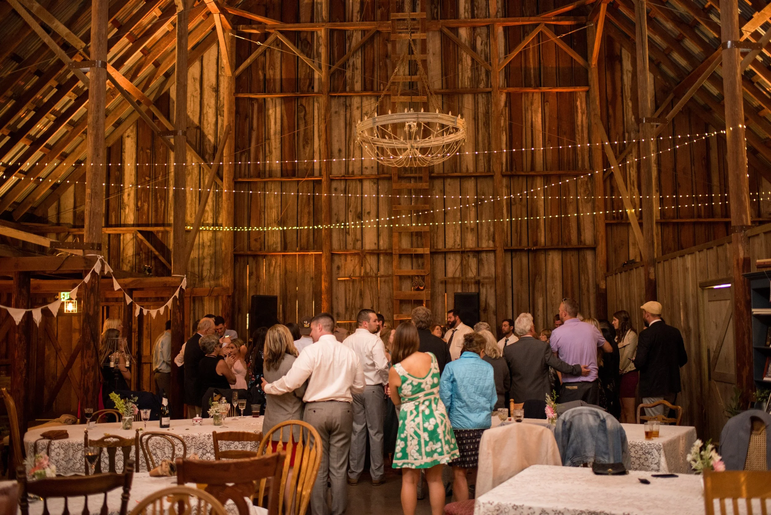 Guests dancing and socializing in a rustic barn decorated with string lights and wedding banners, with tables set for a celebration.