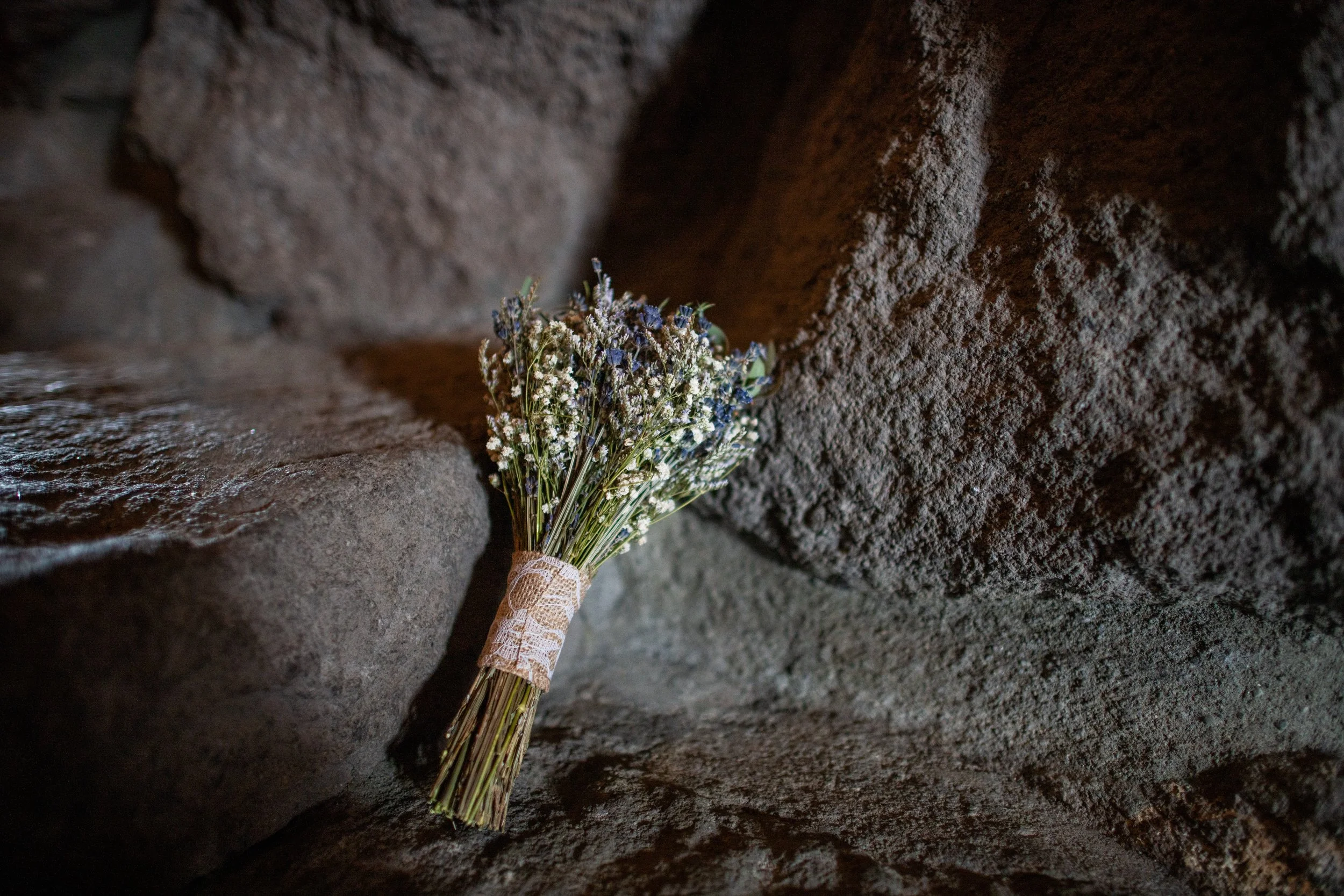 A bouquet of small white and purple flowers wrapped in burlap rests against a dark, rough stone wall.