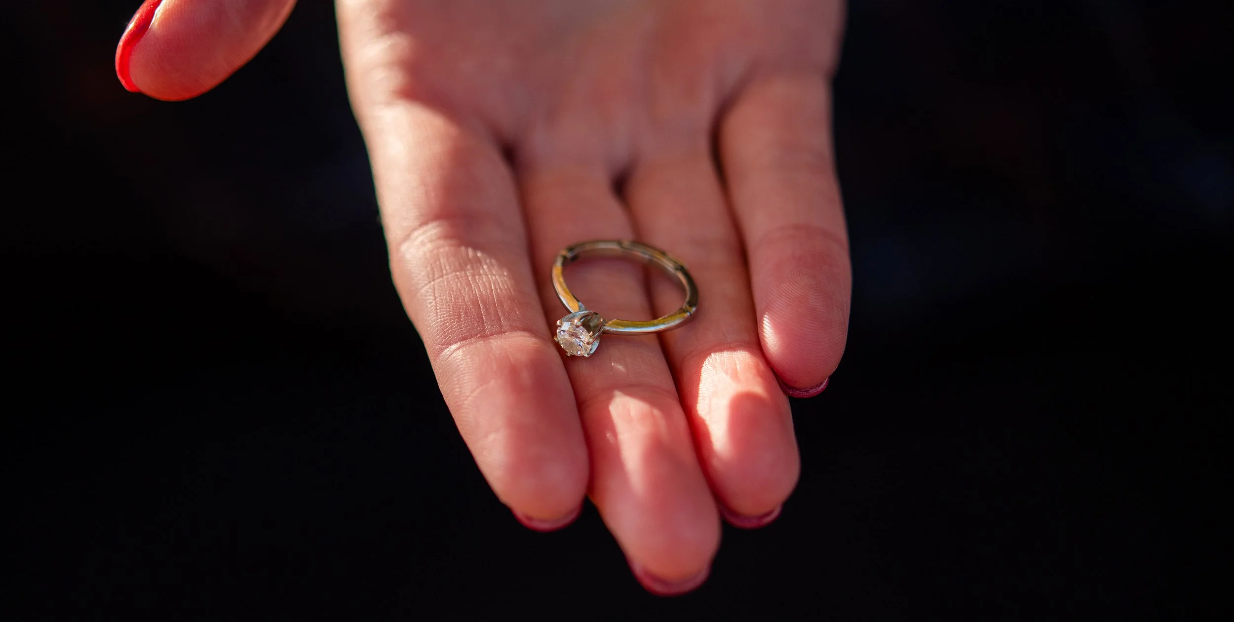 A hand with red-painted nails holding a gold ring with a large, clear gemstone, against a dark background.