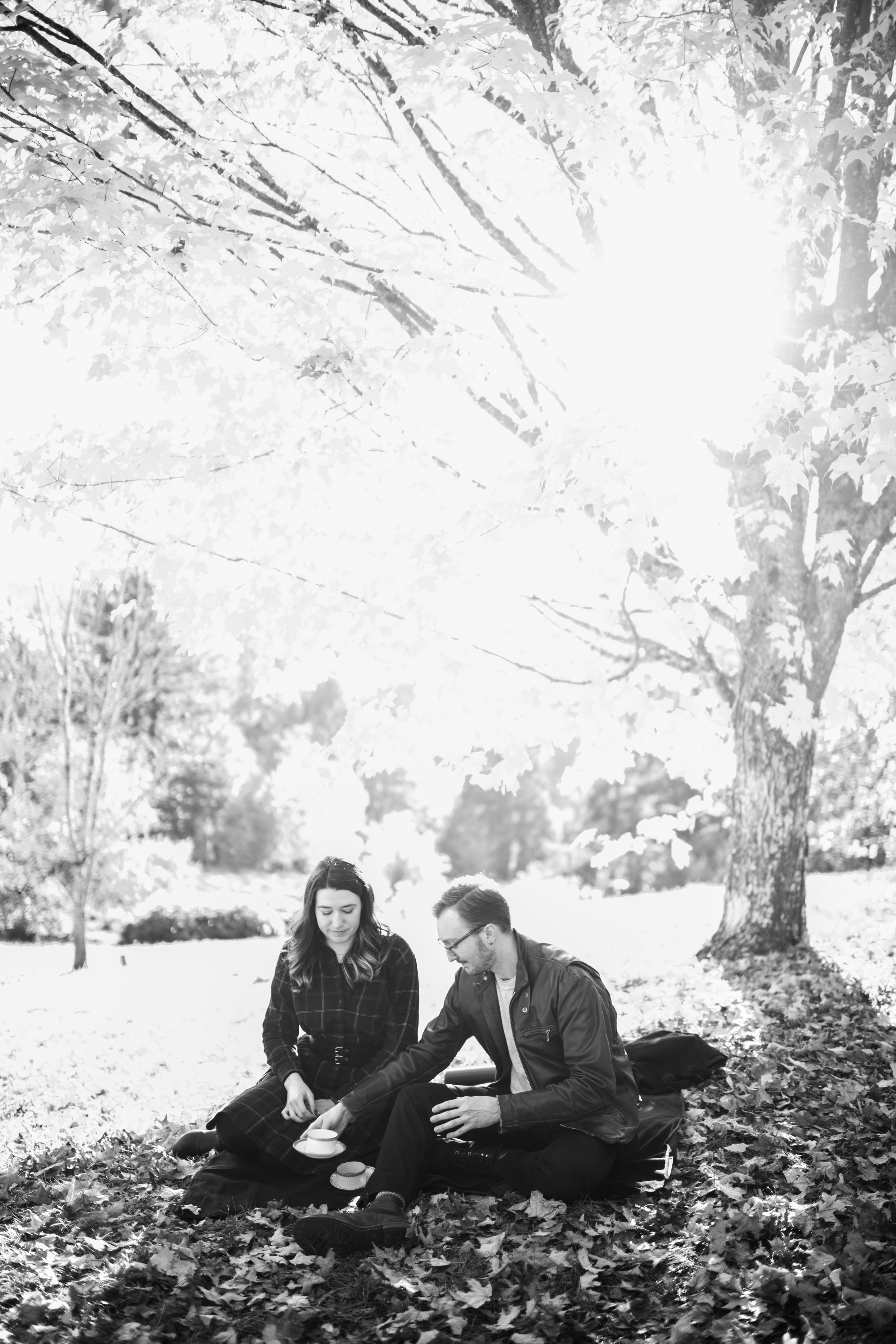 Two young adults, a woman with long hair and a man with glasses, sit on a blanket in a park surrounded by fallen leaves, enjoying a tea or coffee together.