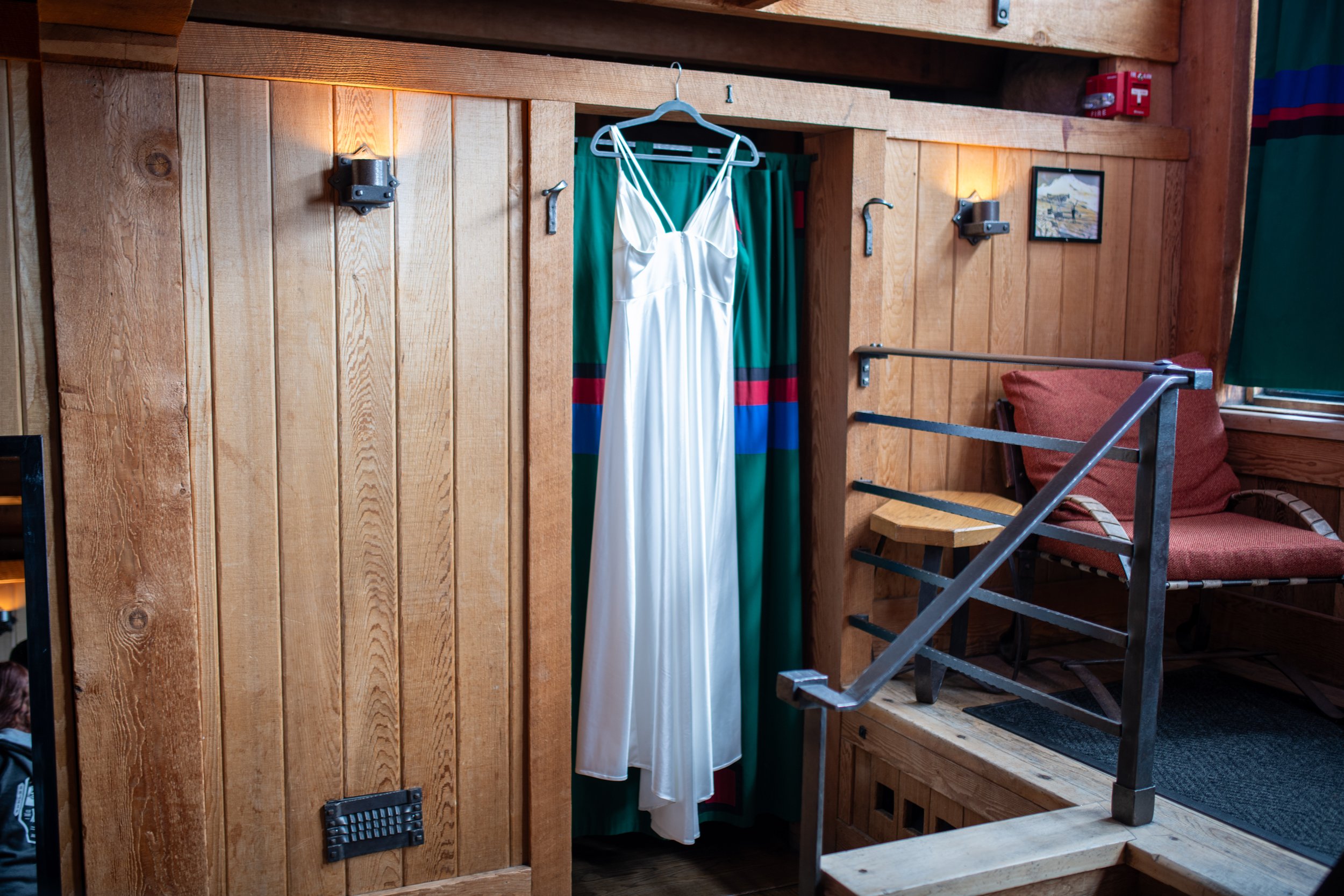 A white dress hanging on a hanger in a wooden room, with a colorful curtain behind it. The room has wooden walls and stairs, with a framed picture and a red fire alarm in the background.