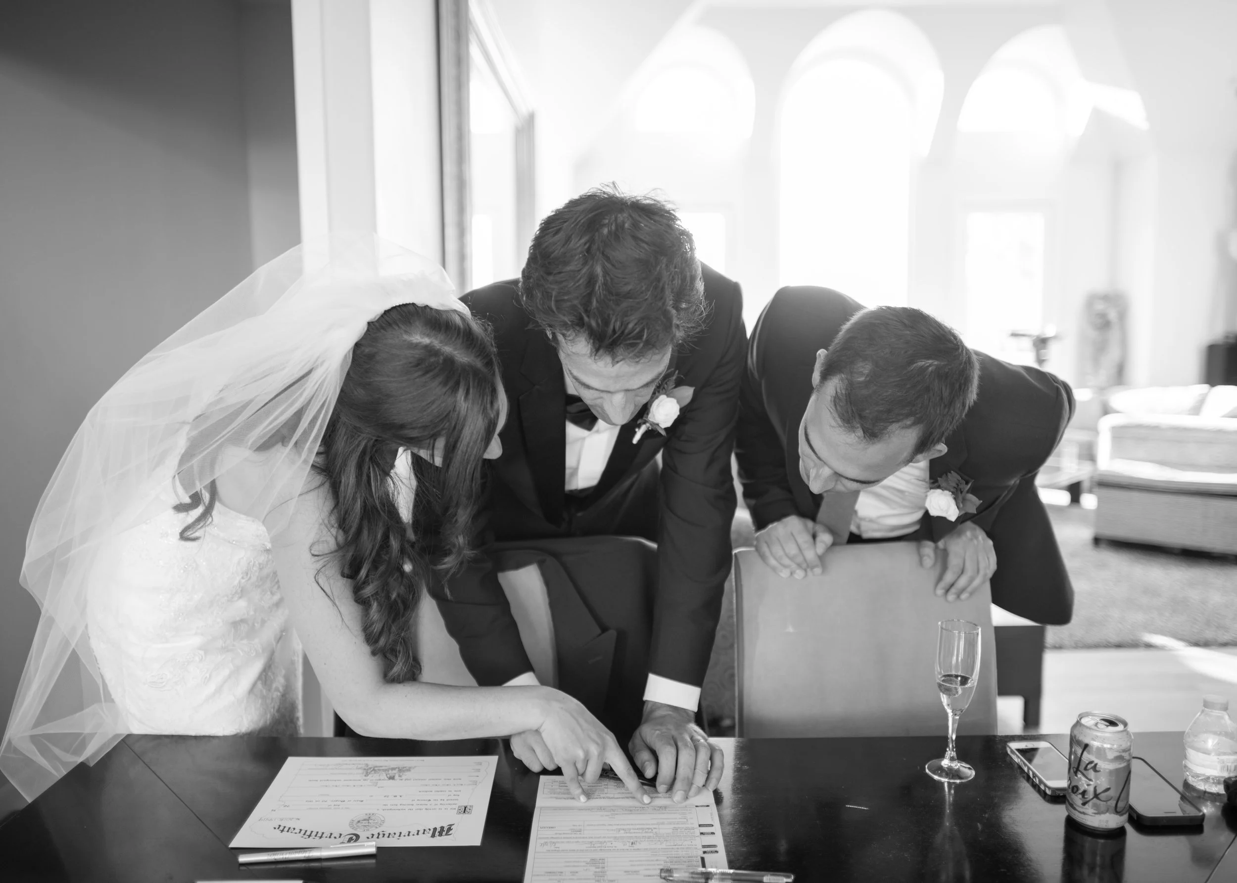 A bride and groom signing a marriage certificate with a officiant during a wedding ceremony, with glasses and water bottles on the table.