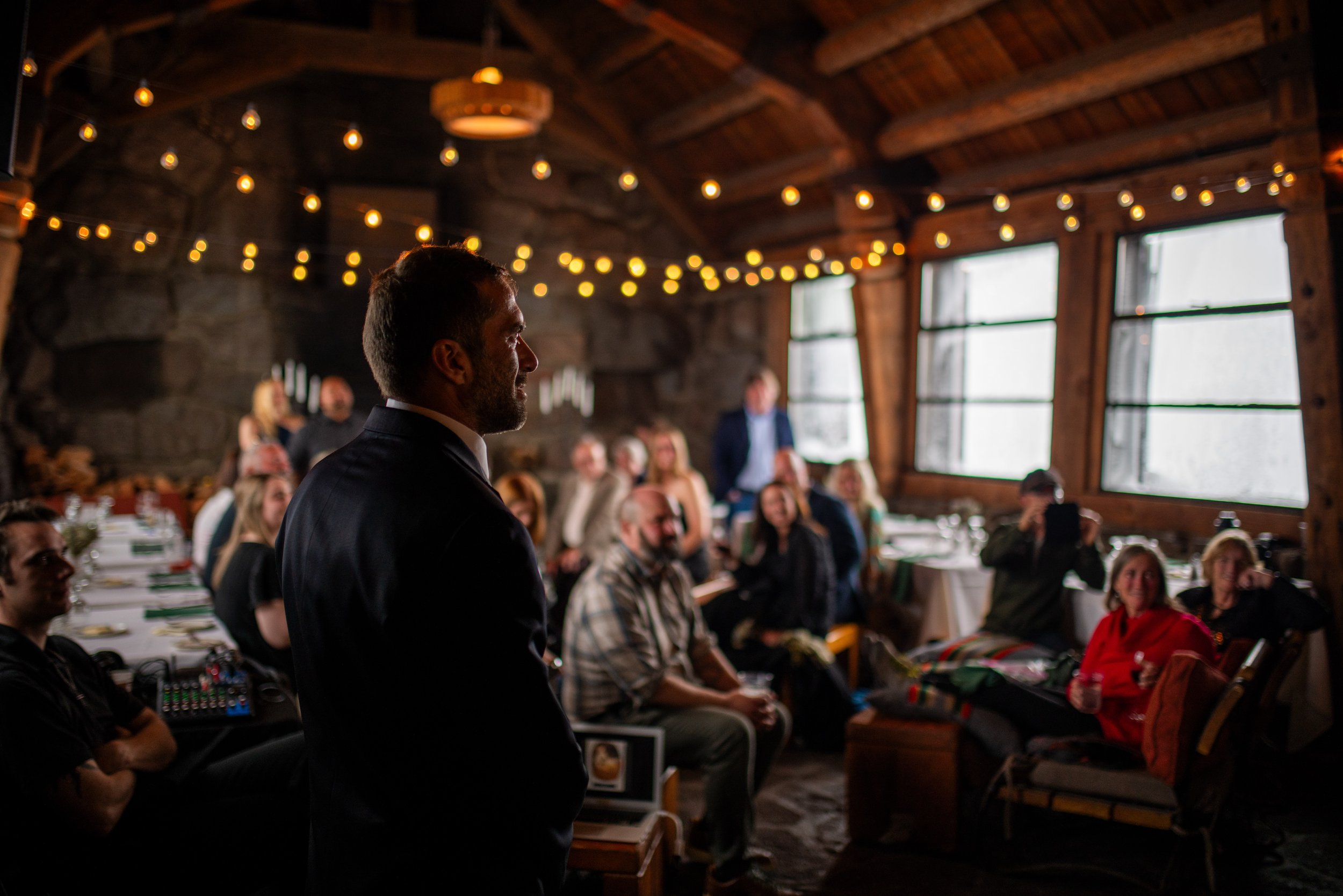 Man in a suit at a large indoor gathering with string lights and wooden beams, people seated at tables.