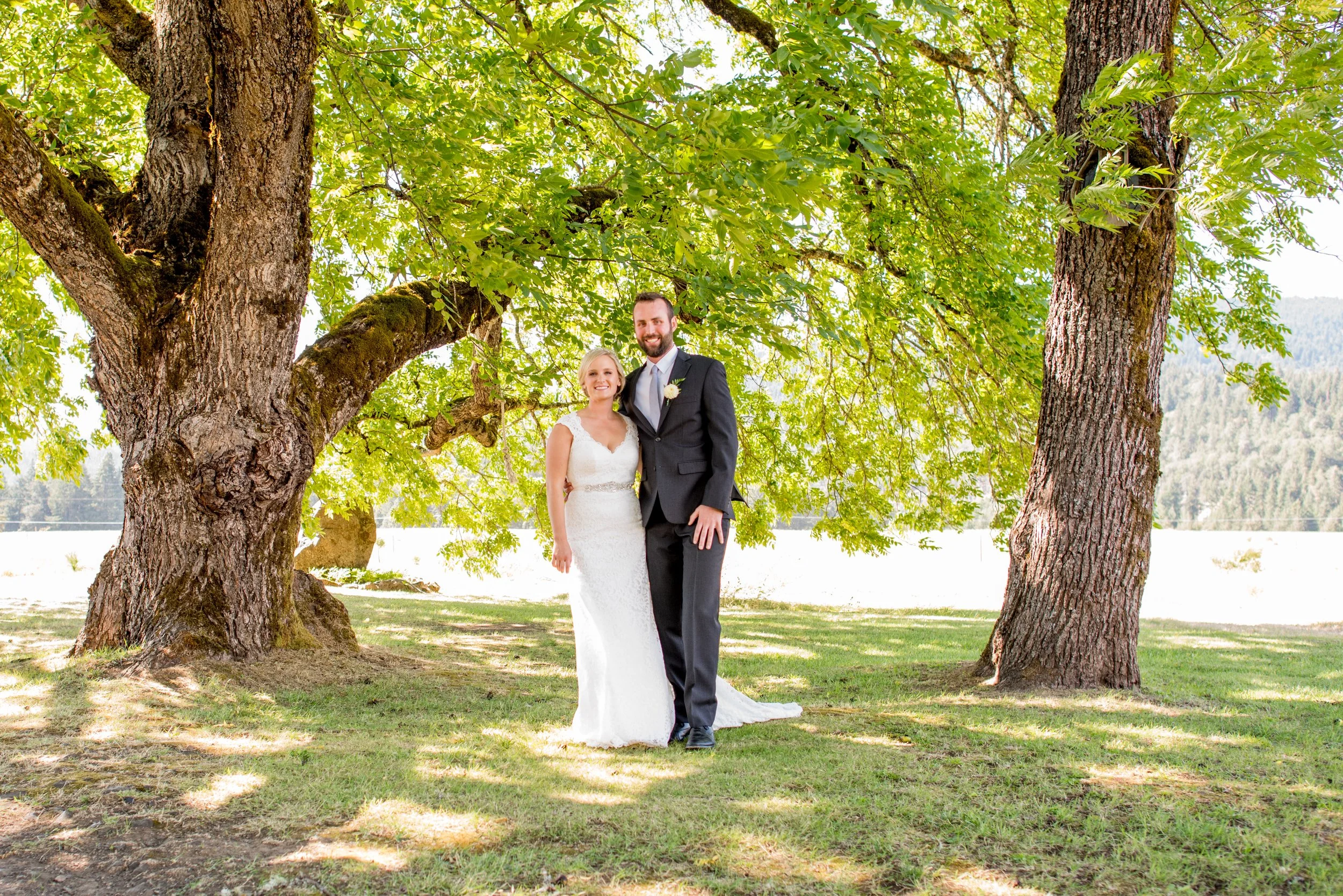 A bride and groom standing together outdoors beneath a large green-leafed tree, smiling, dressed in wedding attire.