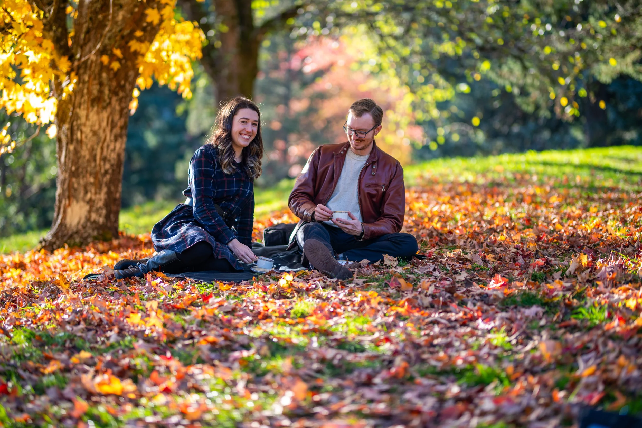 A young man and woman enjoying a picnic on a blanket in a park during autumn, surrounded by fallen colorful leaves and trees with yellow and red leaves in the background.