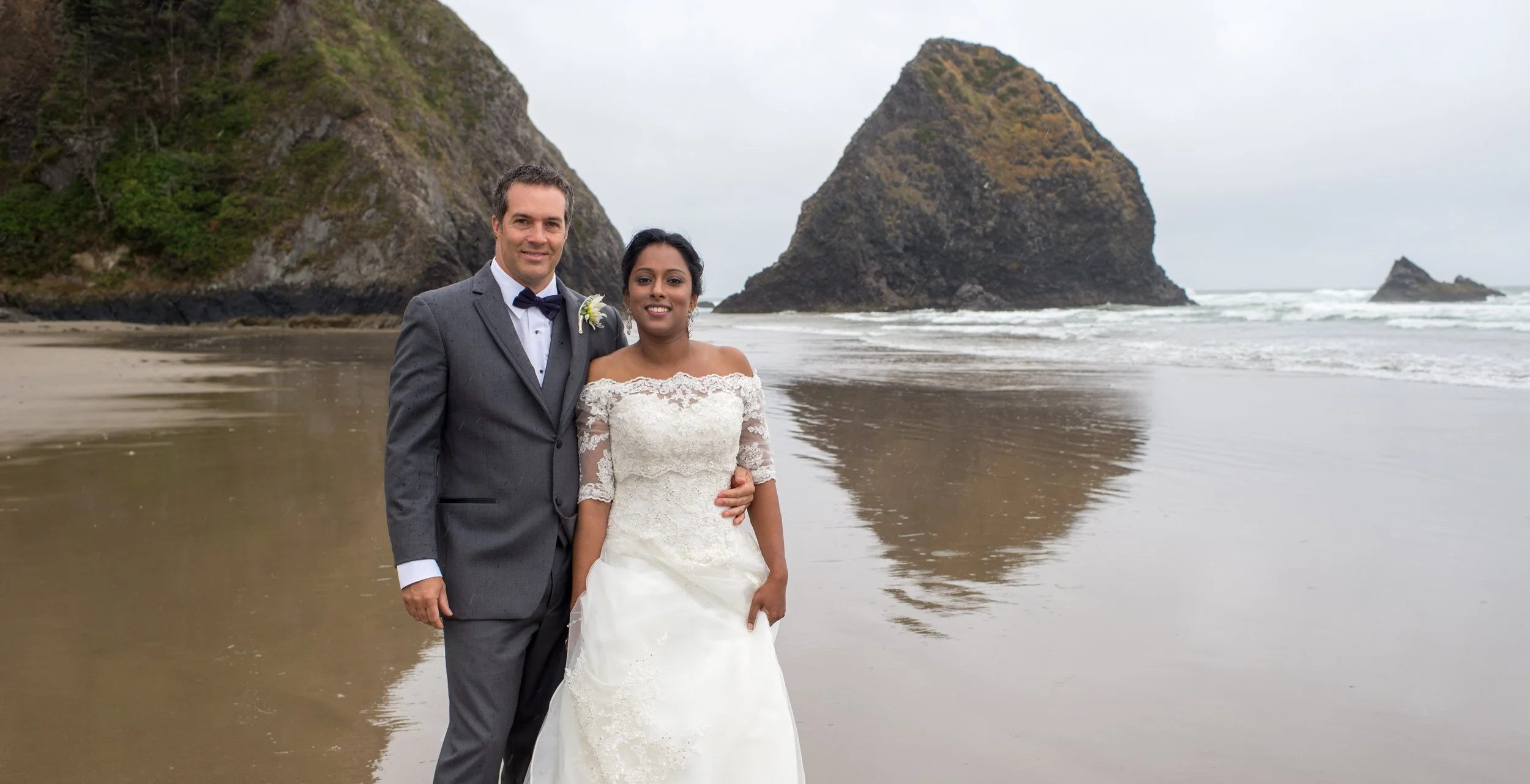 A newlywed couple in wedding attire standing on a beach with large rocks and cliffs in the background, overcast sky.