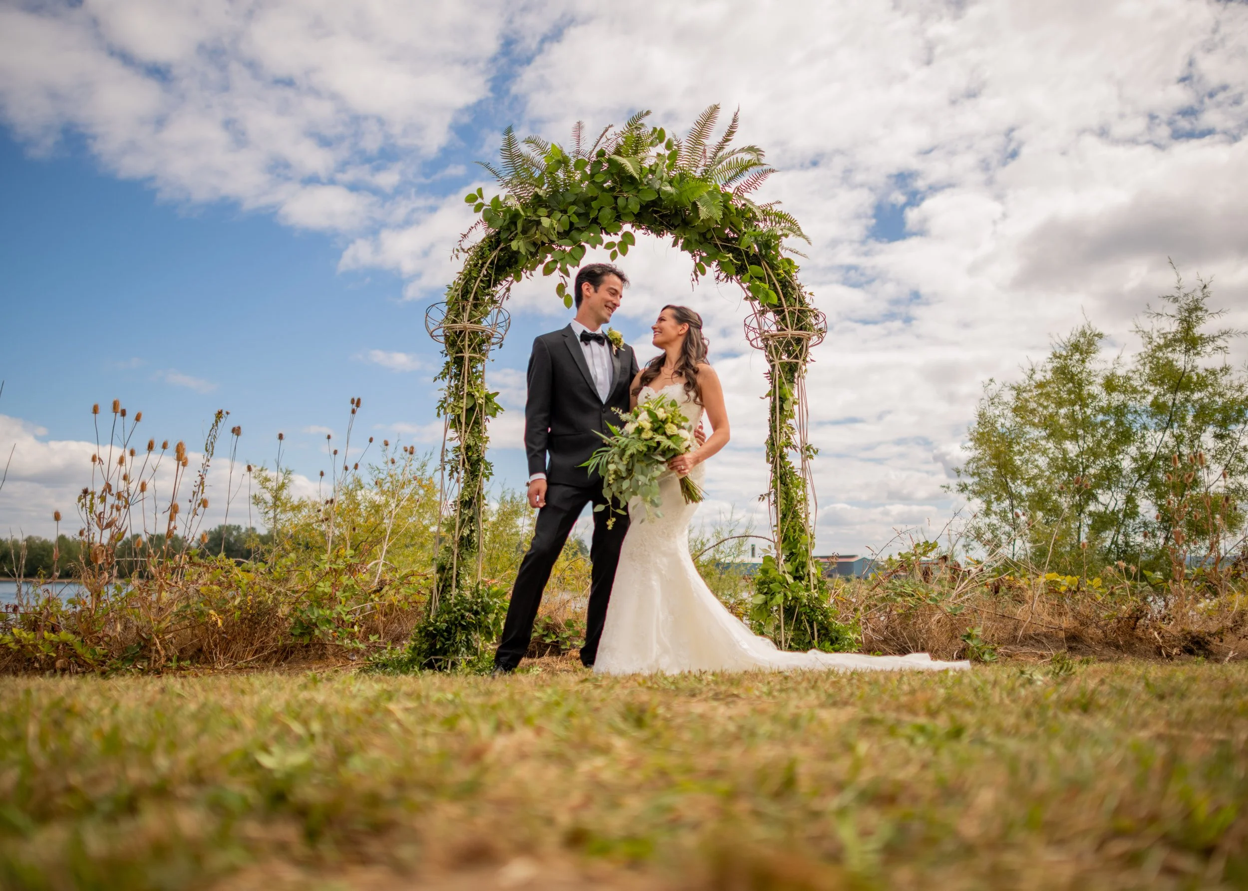 A bride and groom standing under a leafy wedding arch outdoors on a cloudy day.