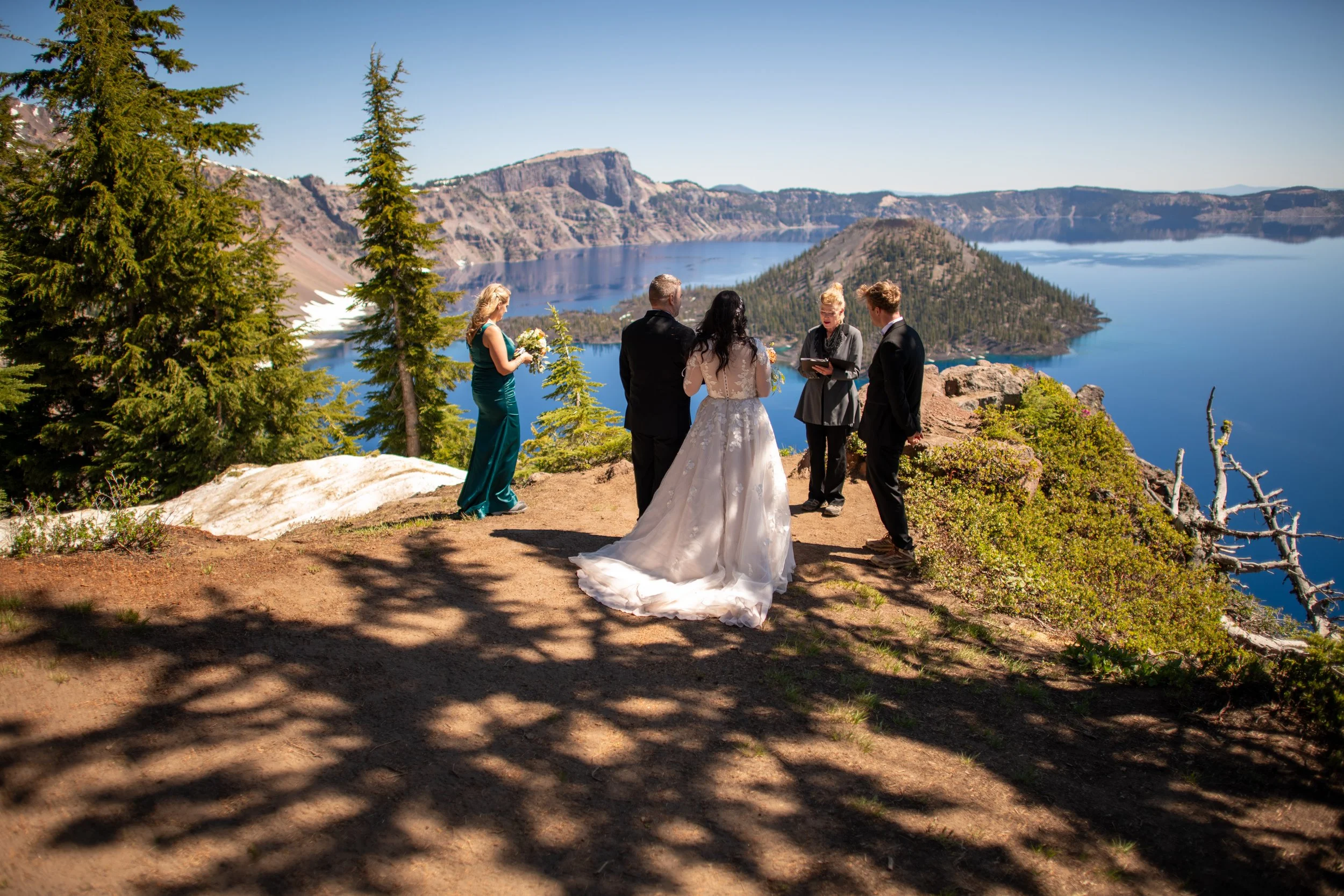 A wedding ceremony taking place outdoors with a scenic view of Crater Lake, surrounded by tall pine trees and mountains in the background.