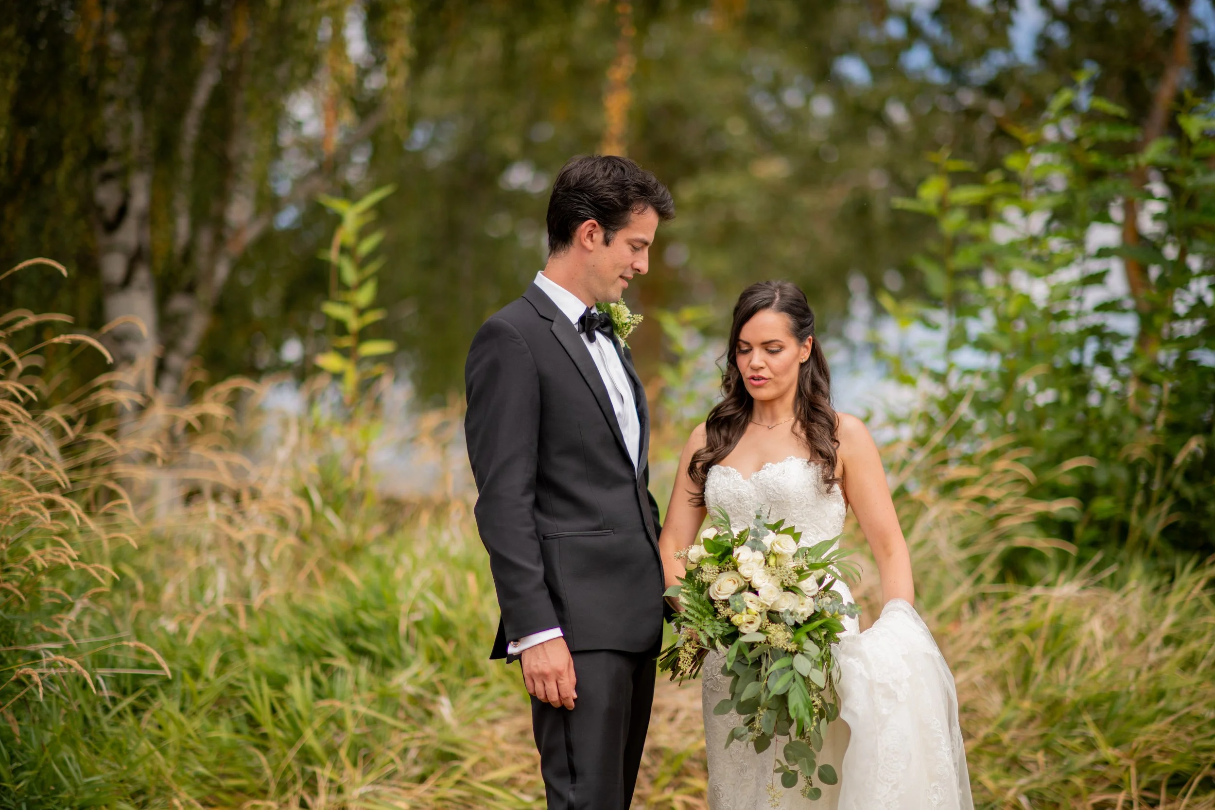 A bride and groom standing outdoors on their wedding day, with the bride holding a bouquet of white roses and greenery, both dressed in formal wedding attire, surrounded by greenery and trees.