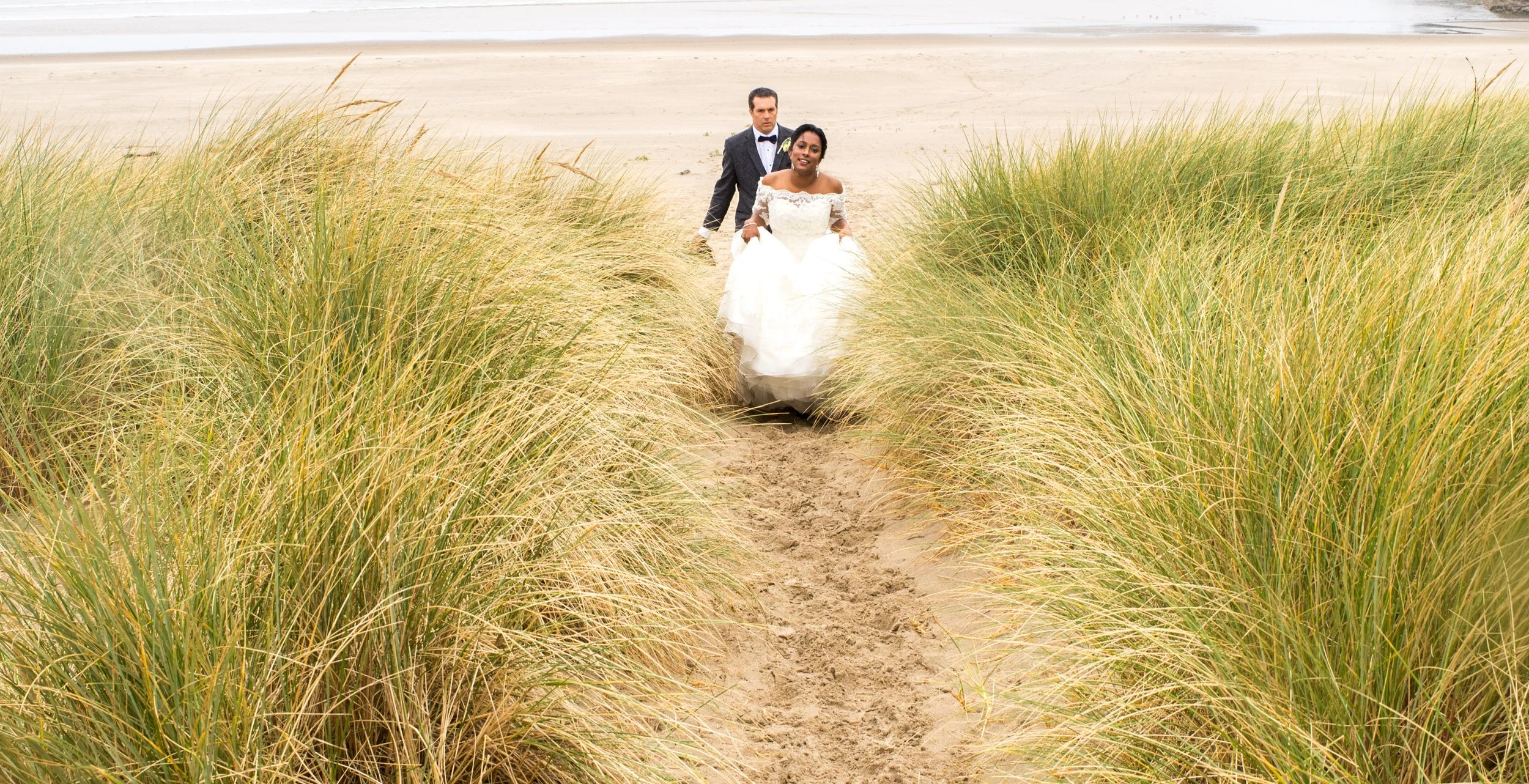A bride and groom walking on a sandy beach through tall grass dunes, with the bride holding her wedding dress and the groom dressed in a suit with a bow tie.