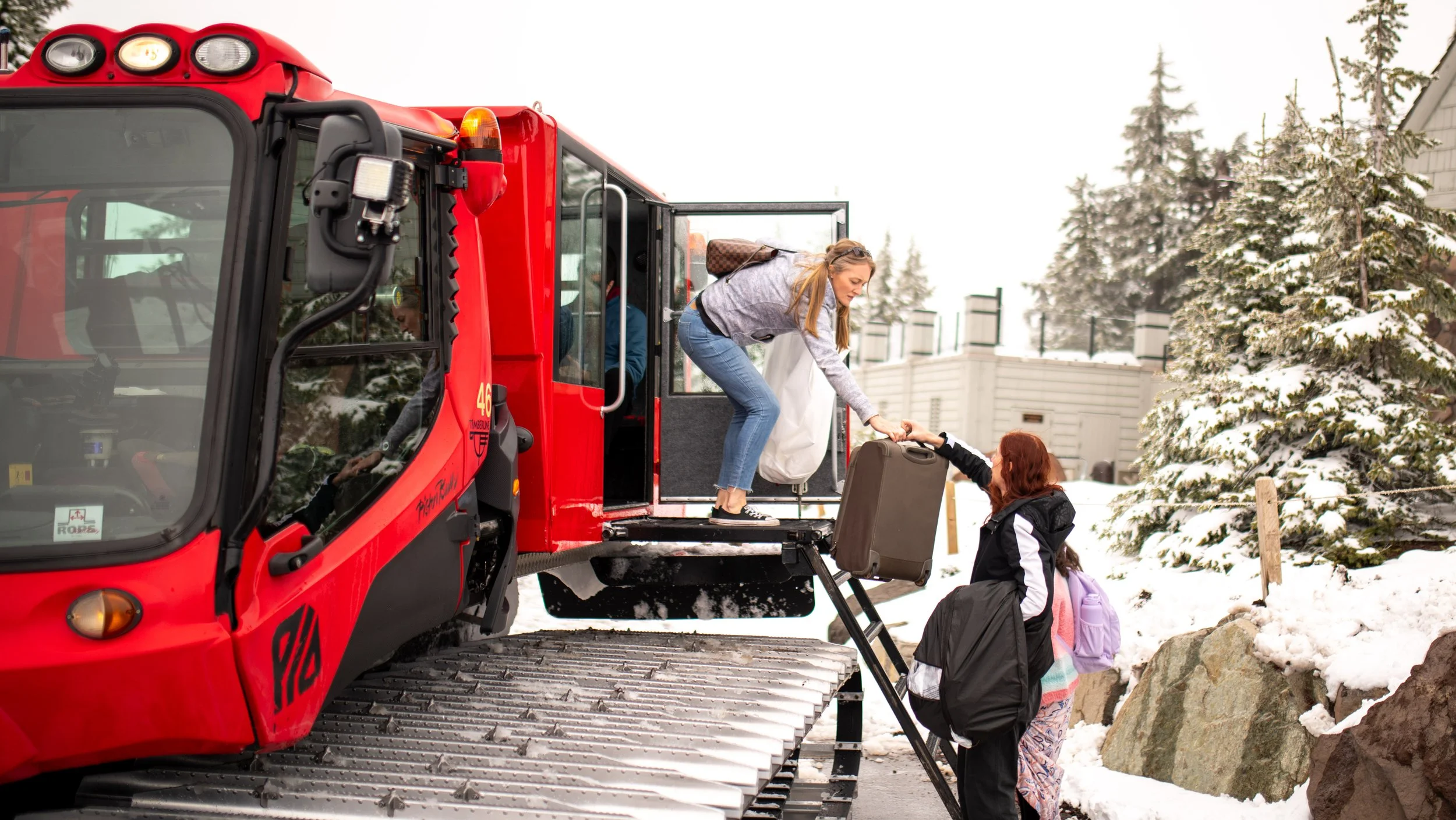 A woman boarding a red snowcat vehicle with passengers, helping a young girl with a backpack, in a snowy outdoor setting with trees and rocks.