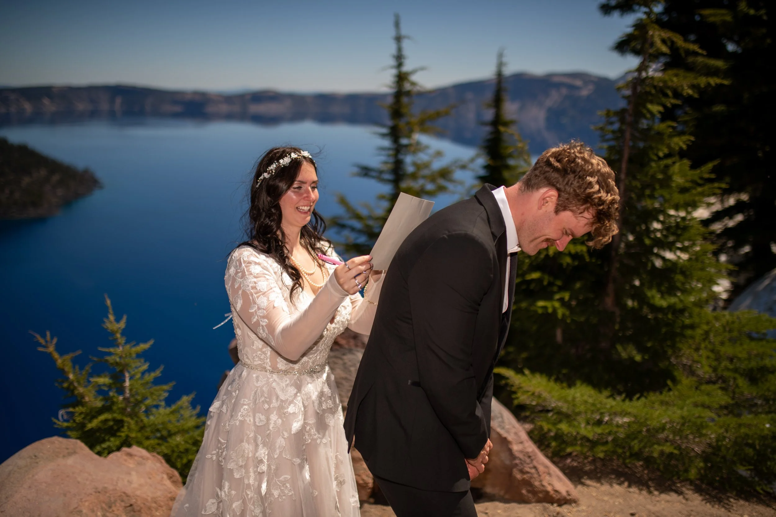 A bride in a wedding dress holds a paper and pen behind a groom in a suit, who is leaning forward with his hands clasped, in an outdoor setting near a large lake with mountains and trees in the background.