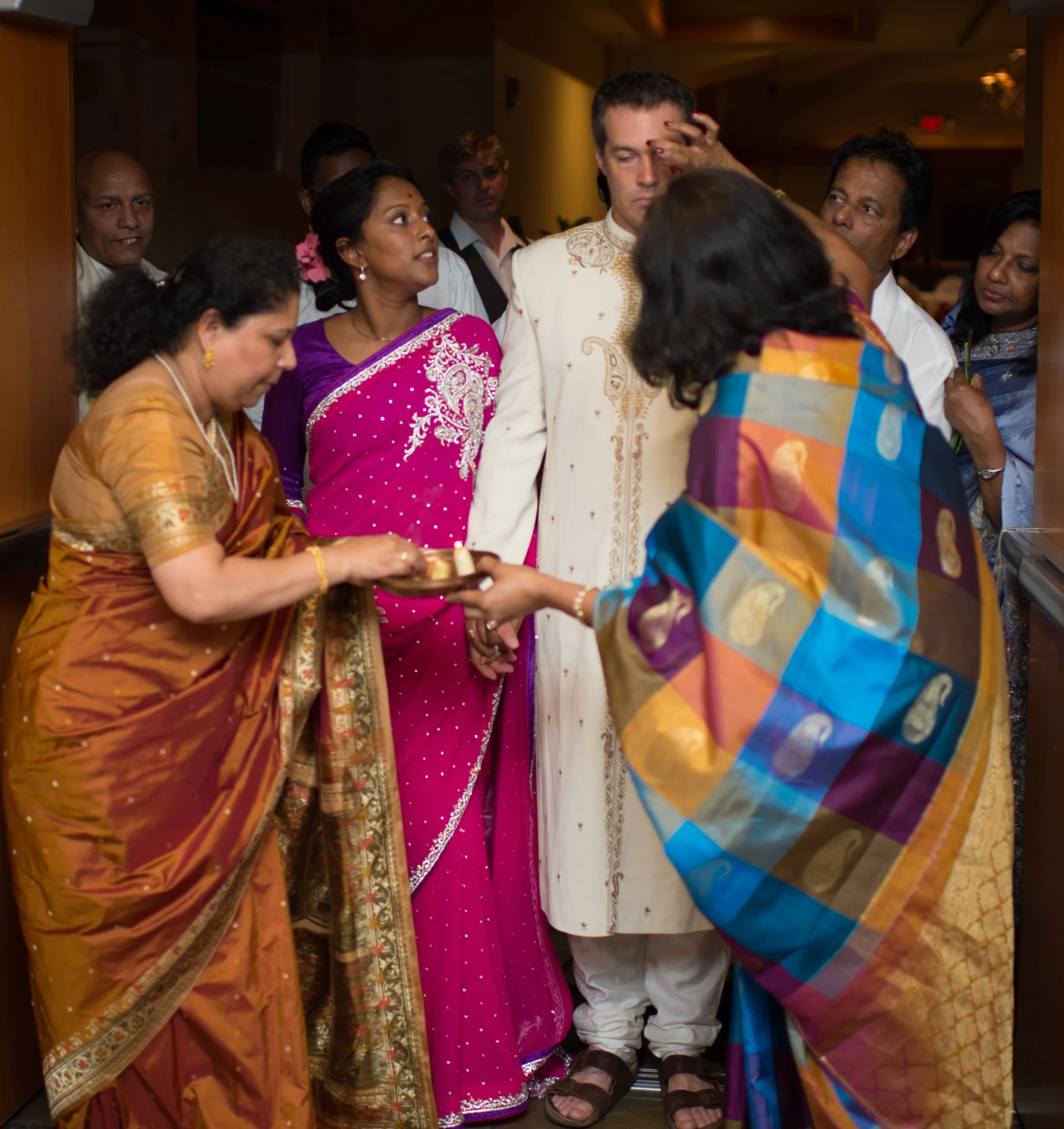 Indian woman in orange sari holding a tray, woman in pink sari with flowers in her hair, man with a hand on his forehead, woman in multicolored sari, and other people gathered in an indoor setting for a traditional ceremony.
