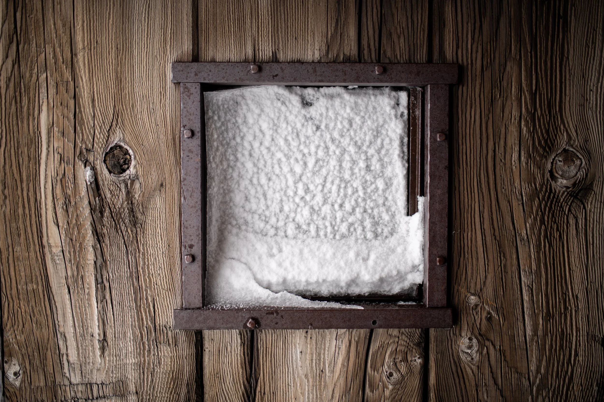 A metal vent cover with snow on it, mounted on a wooden wall.