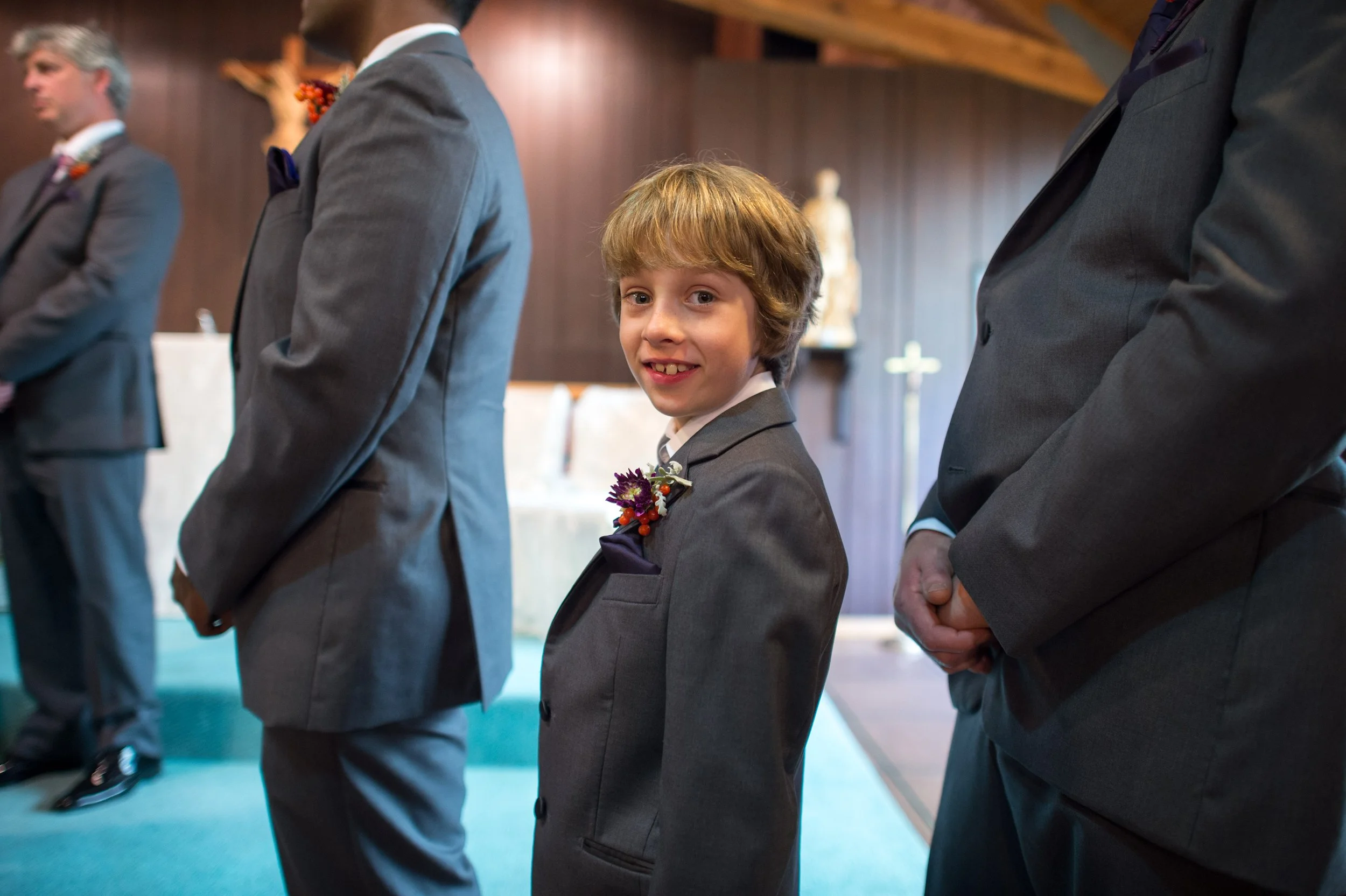 Young boy and two men dressed in suits lined up in a wedding ceremony, standing with hands clasped in front of them.