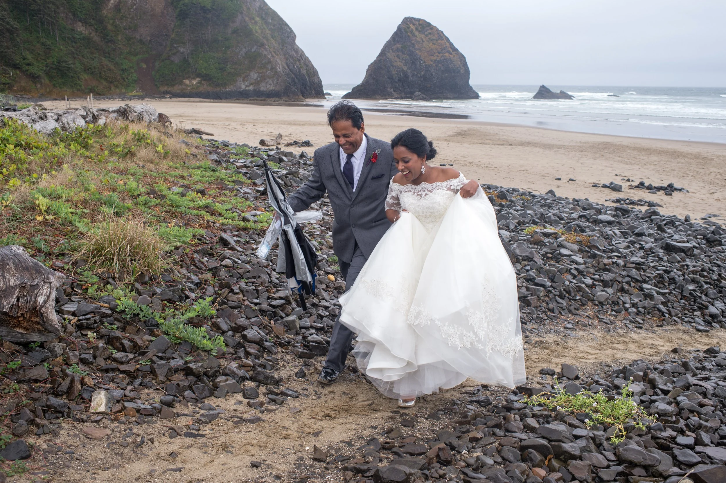 A couple dressed in wedding attire walking along a rocky beach with large sea stacks in the background.