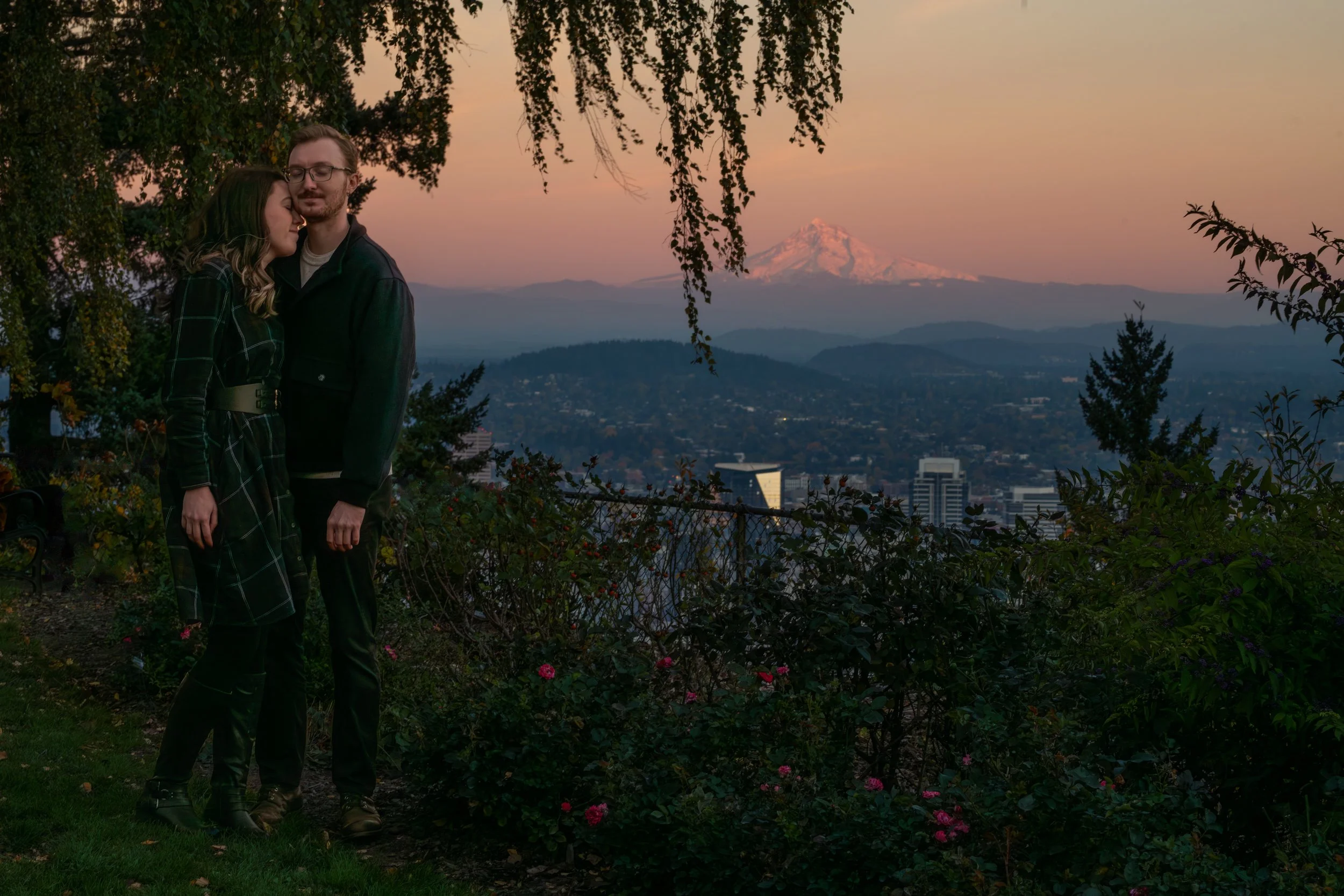 A couple stands close together in a garden at sunset, with Mount Hood in the background and cityscape below.
