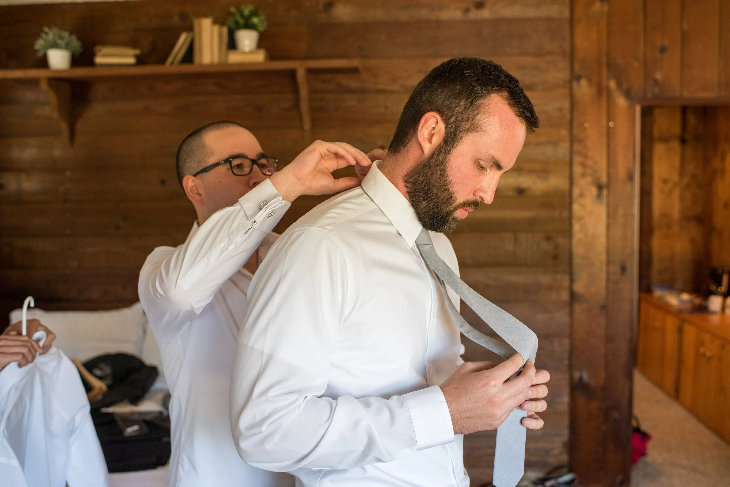 A man with a beard dressed in a white shirt is being assisted by another man in a white shirt, who is adjusting his tie in a room with wooden walls and shelves holding books and potted plants.