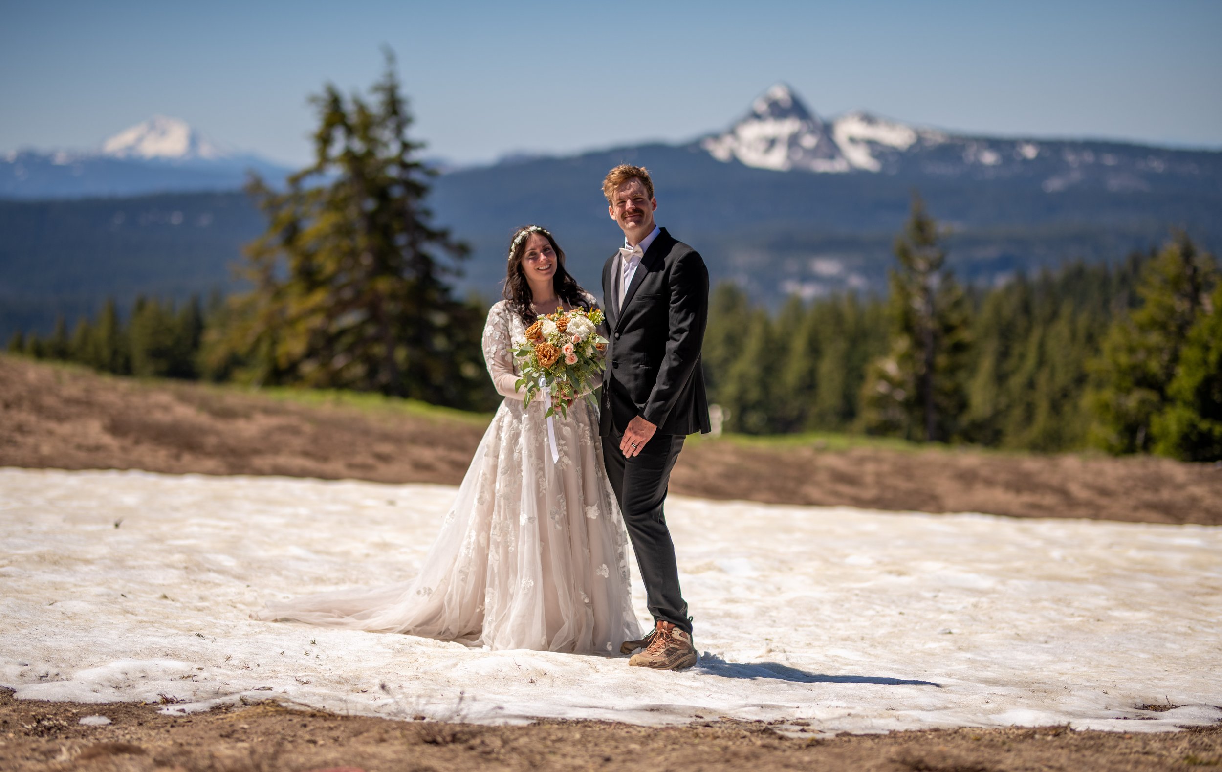 A bride and groom standing on a dirt area in an outdoor setting with mountains and trees in the background. The bride is wearing a wedding dress and holding a bouquet, while the groom is in a tuxedo.