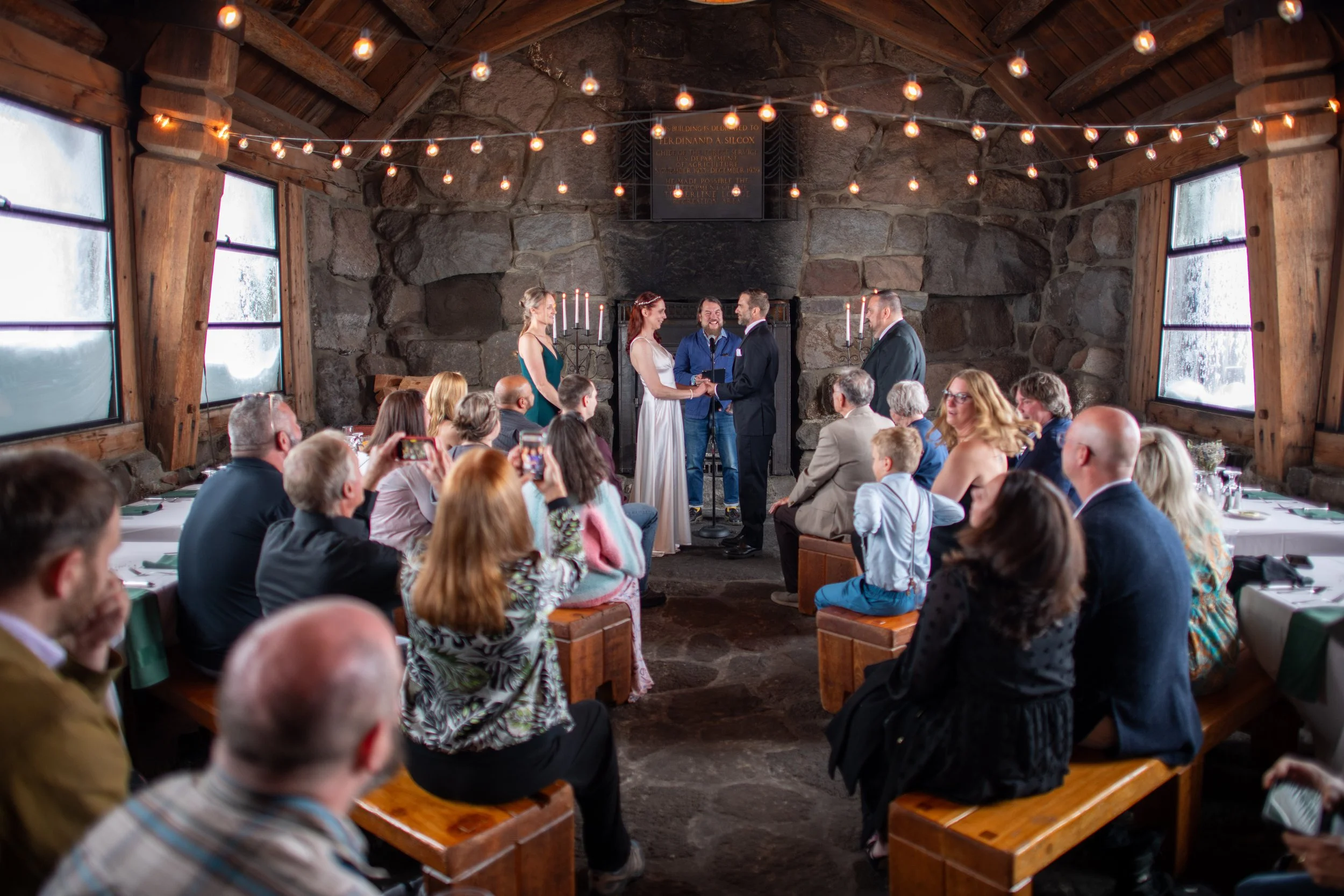 A couple is getting married inside a rustic stone and wood venue, standing in front of an officiant. Guests are seated at long tables, watching and taking pictures. String lights are hanging from the ceiling, and candles are on the fireplace behind t