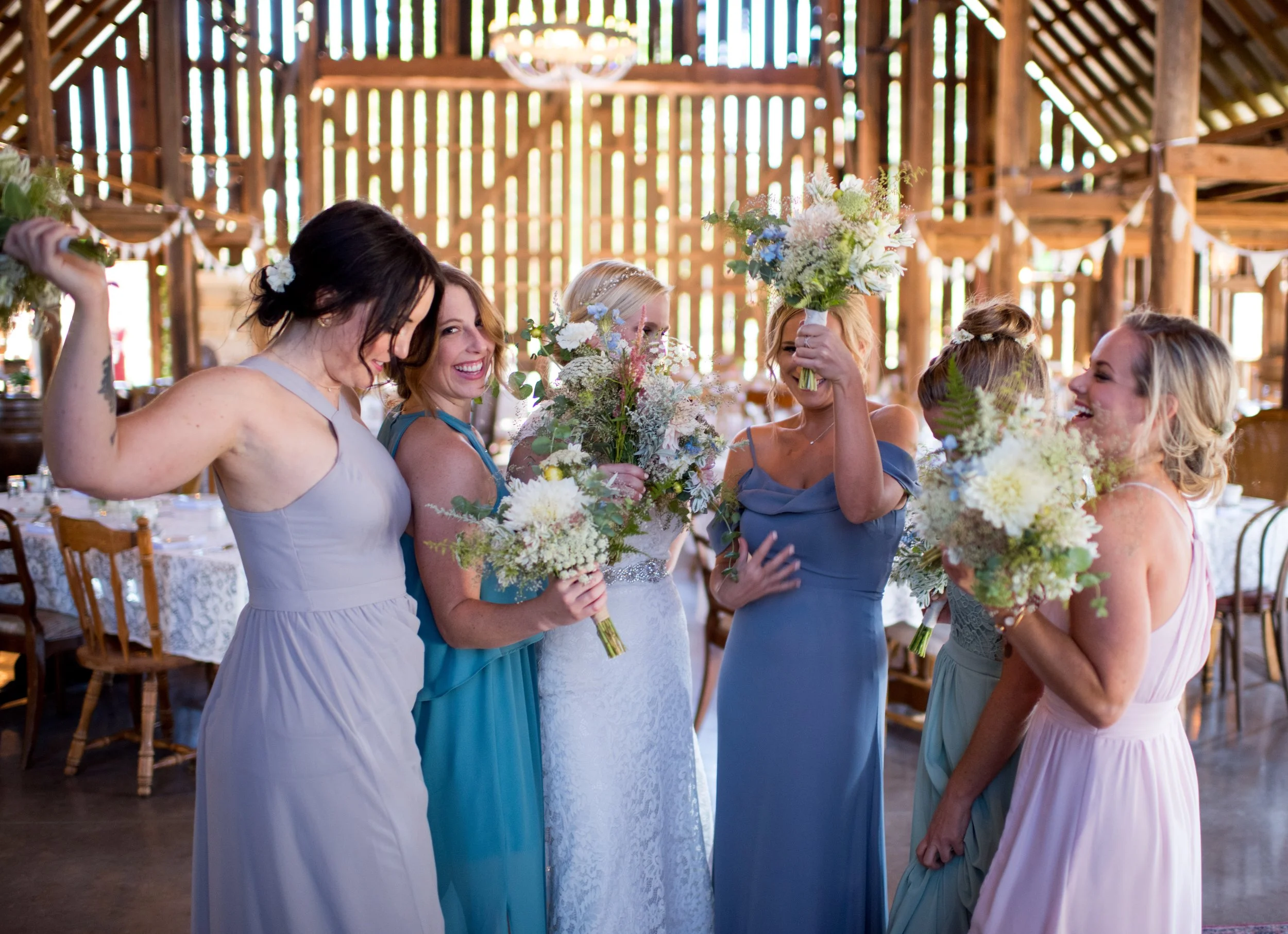 Group of women in dresses holding bouquets of flowers at a wedding or celebration in a rustic barn setting.