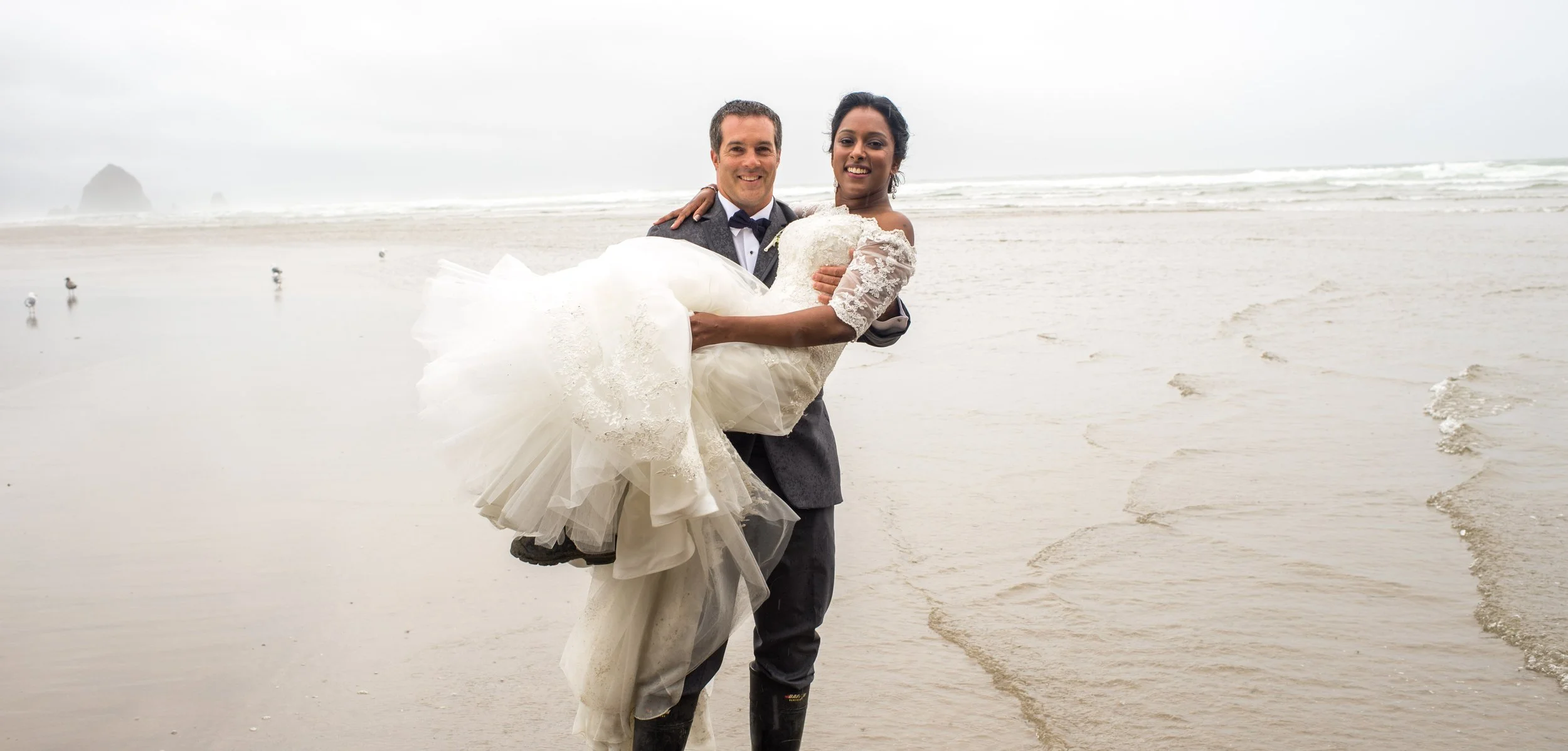 A groom carrying a bride in a white wedding dress on a beach with waves and seagulls in the background.