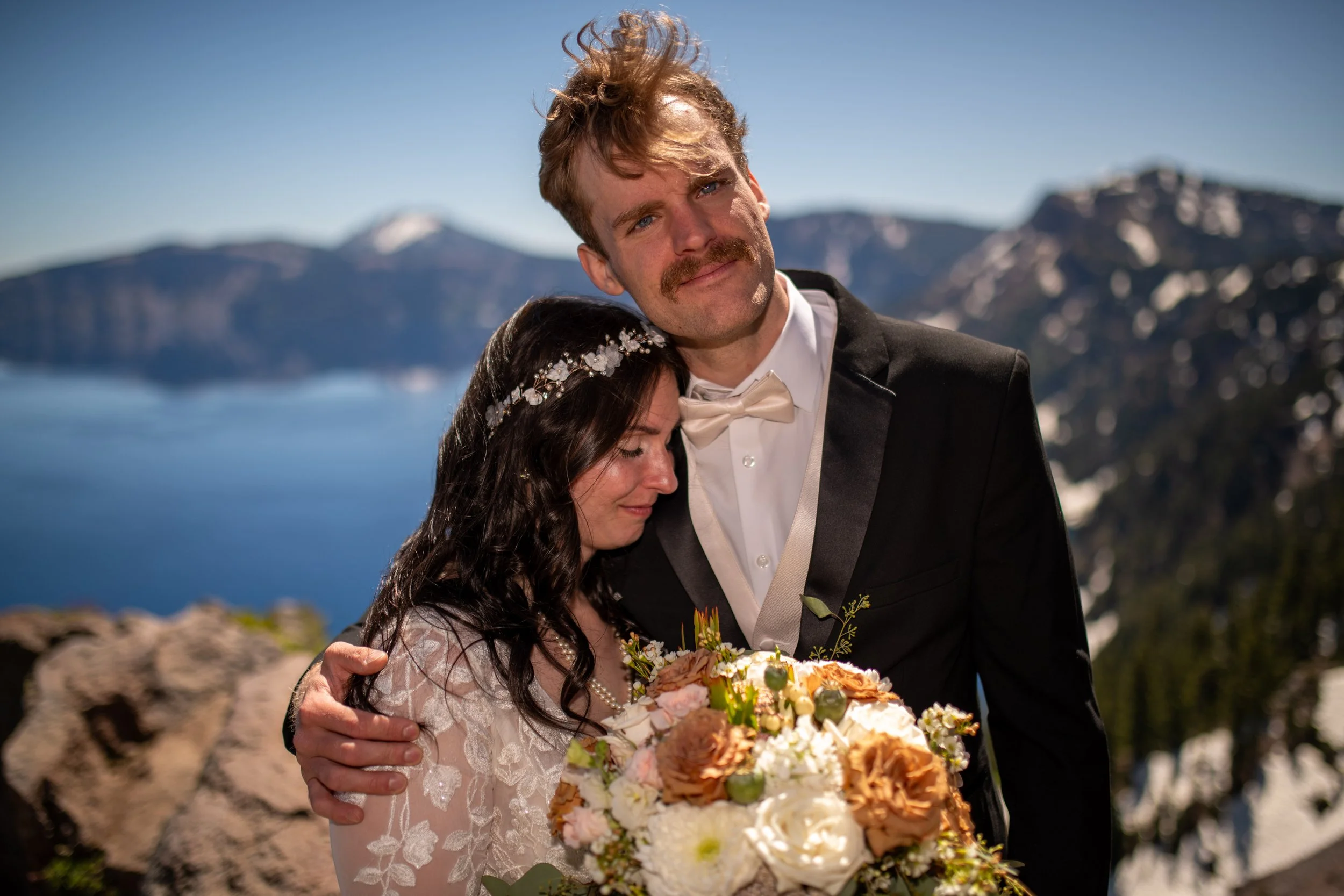 A groom in a tuxedo with a bow tie and a bride with a floral hairpiece and white dress holding a large bouquet of flowers, with a mountain and lake background.