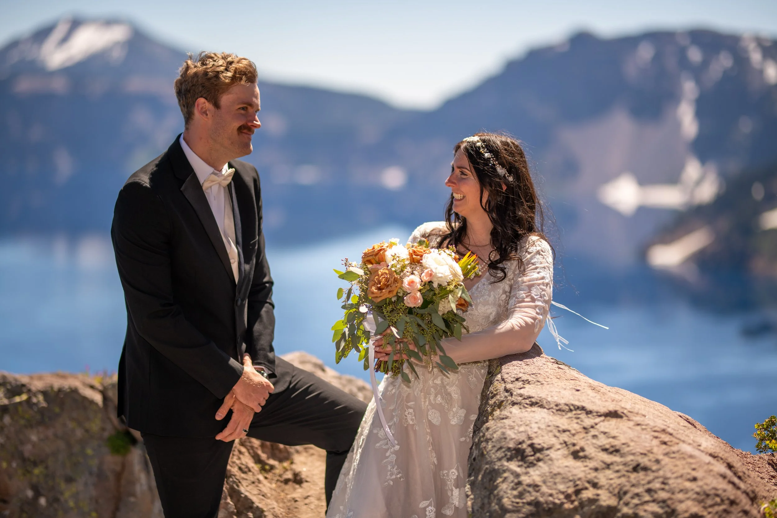 A bride and groom exchanging vows outdoors near a mountain lake with snow-capped peaks in the background, the bride holding a large bouquet of flowers.