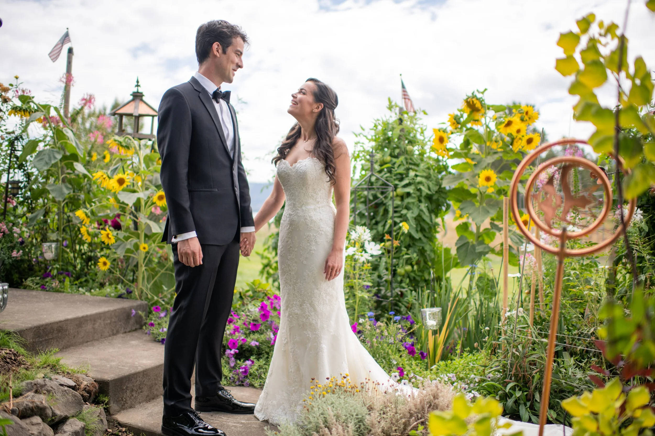A bride and groom holding hands and looking at each other in a garden setting during their wedding ceremony, surrounded by colorful flowers and greenery.