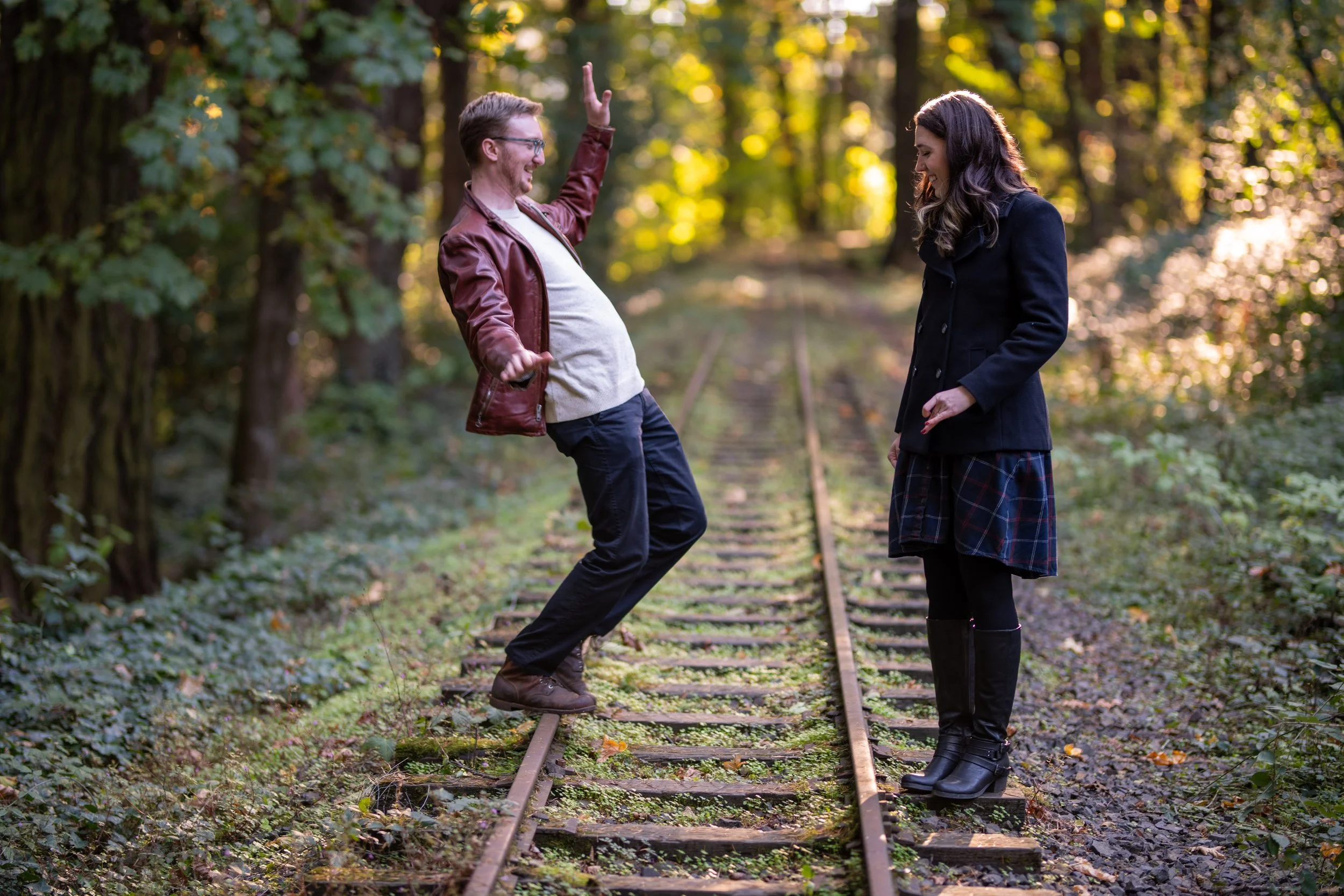 A man and woman are standing on a set of train tracks in a wooded area, smiling and sharing a joyful moment. The man is wearing glasses, a brown leather jacket, and jeans, while the woman is dressed in a dark coat, plaid skirt, and is wearing boots. 